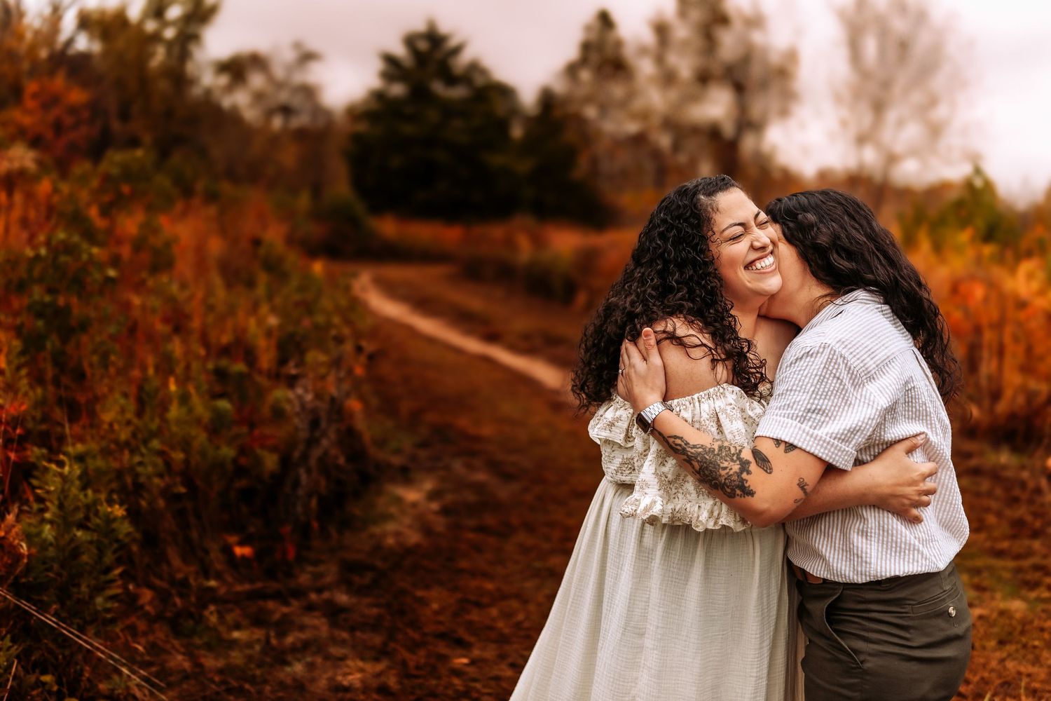 Two women embracing and laughing on an autumn trail surrounded by vibrant orange and red foliage.