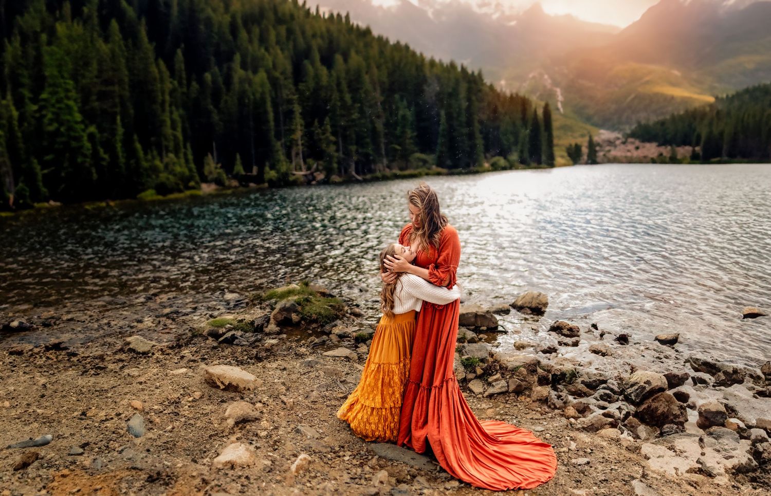Woman in orange dress standing by a mountain lake surrounded by pine trees and rocky shores.
