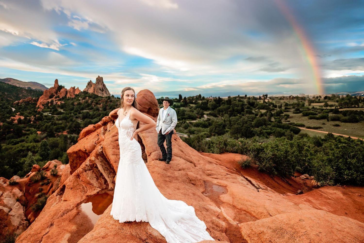 Bride and groom pose on red rocks at Garden of the Gods with a rainbow arching over Colorado's stunning landscape.