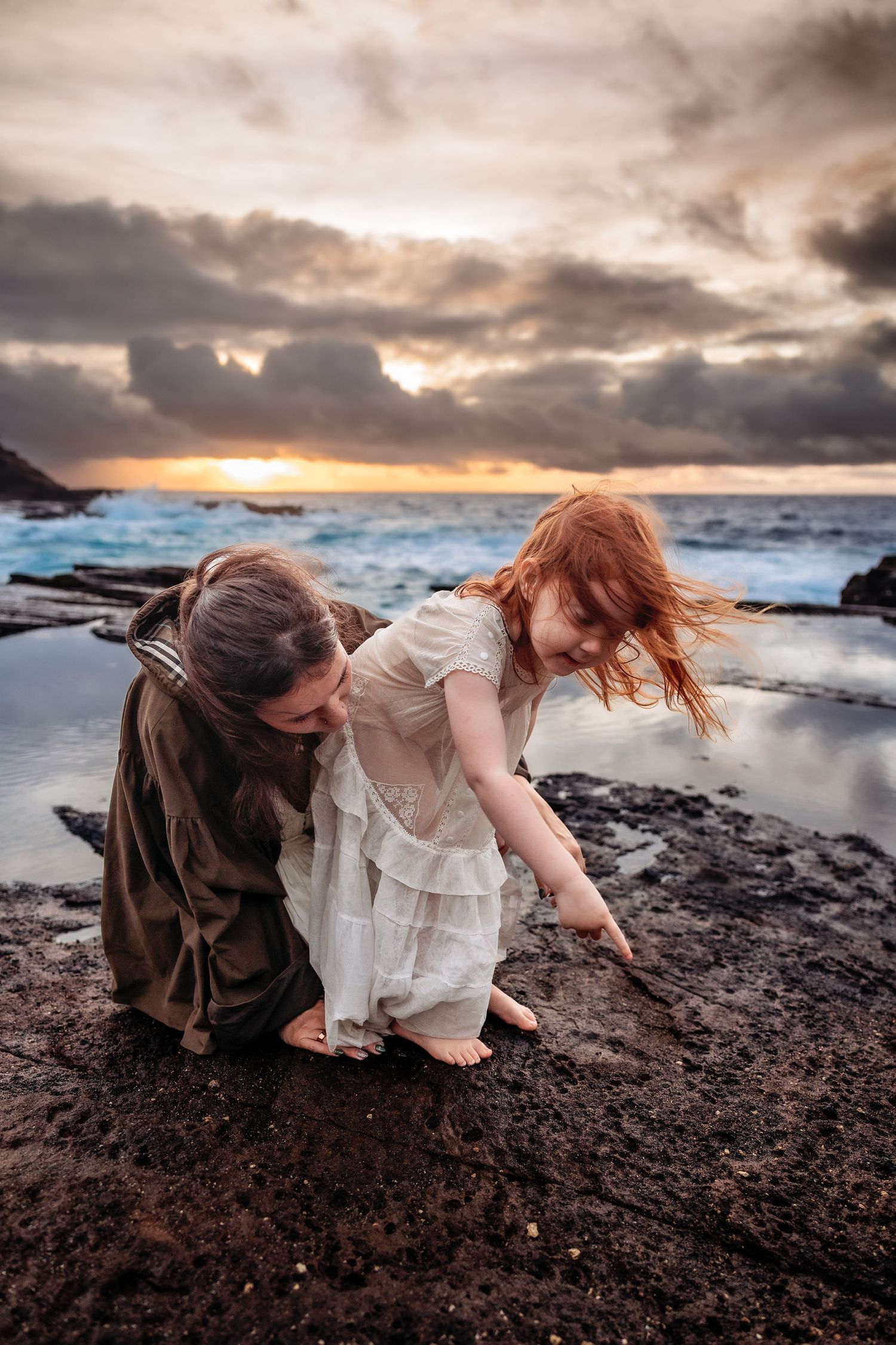 Woman and red-haired girl explore rocky shoreline at sunset with dramatic cloudy sky and ocean waves behind them.