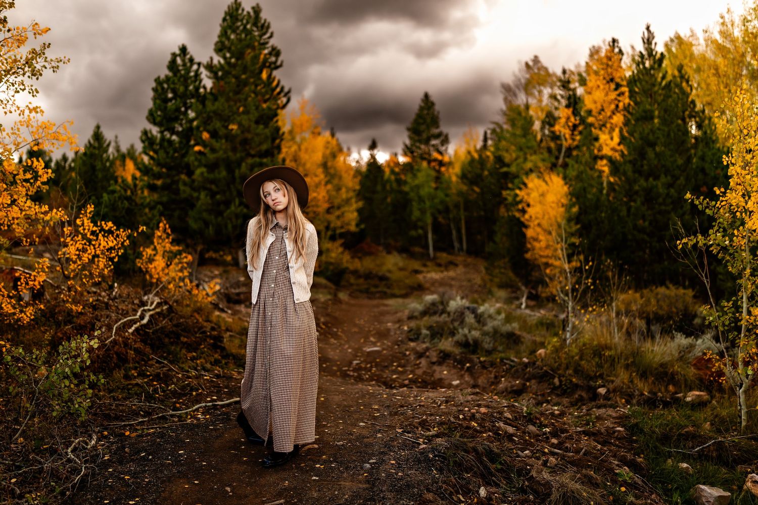 Young woman in maxi dress stands on autumn trail surrounded by golden aspens and evergreens under dramatic stormy sky.