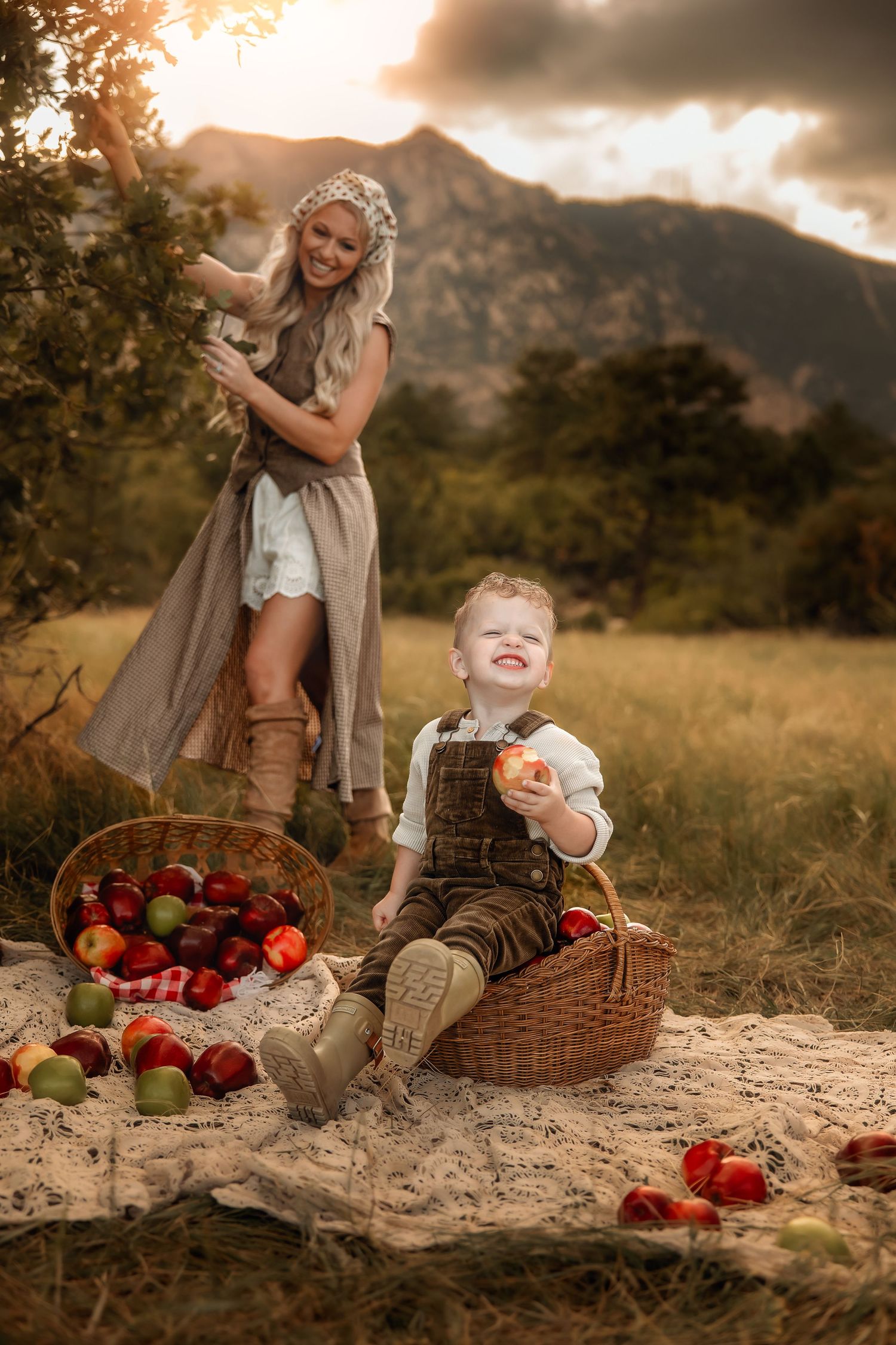A smiling woman picks apples from a tree while a laughing toddler in overalls sits on a blanket with baskets of red apples near mountains.