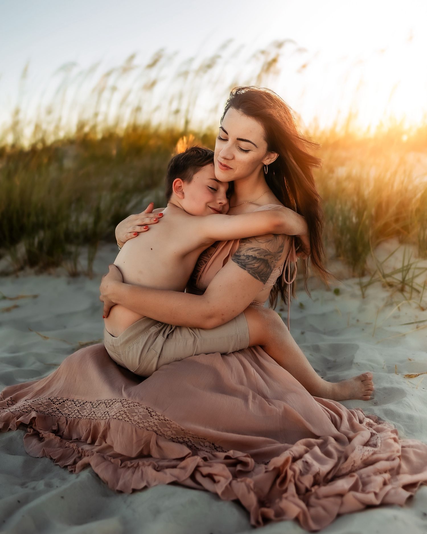 A mother tenderly embraces her child on a sandy beach at golden sunset, wearing a flowing dusty rose dress.