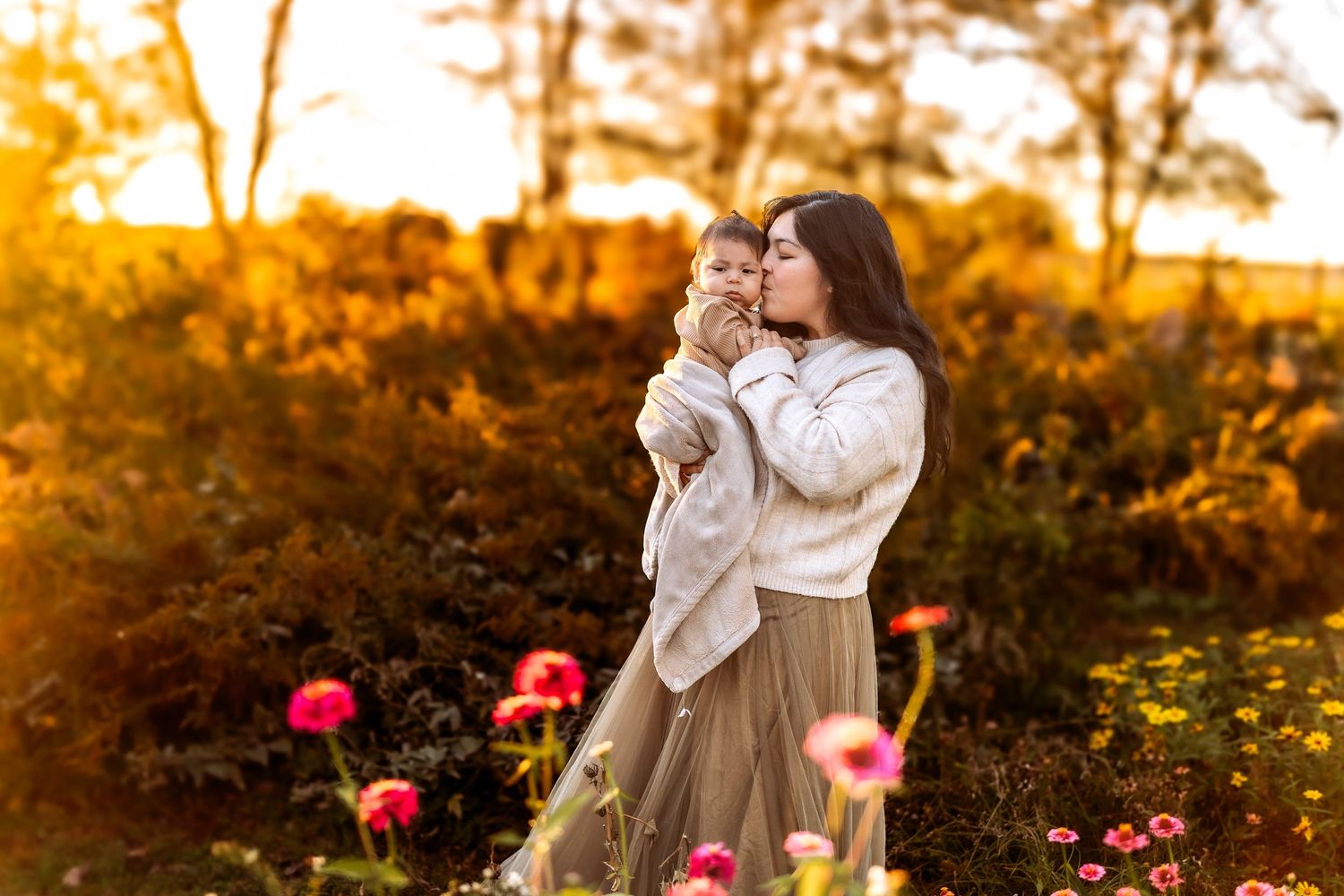 Mother kissing her baby in a golden sunset flower garden, warm autumn light glowing behind them.