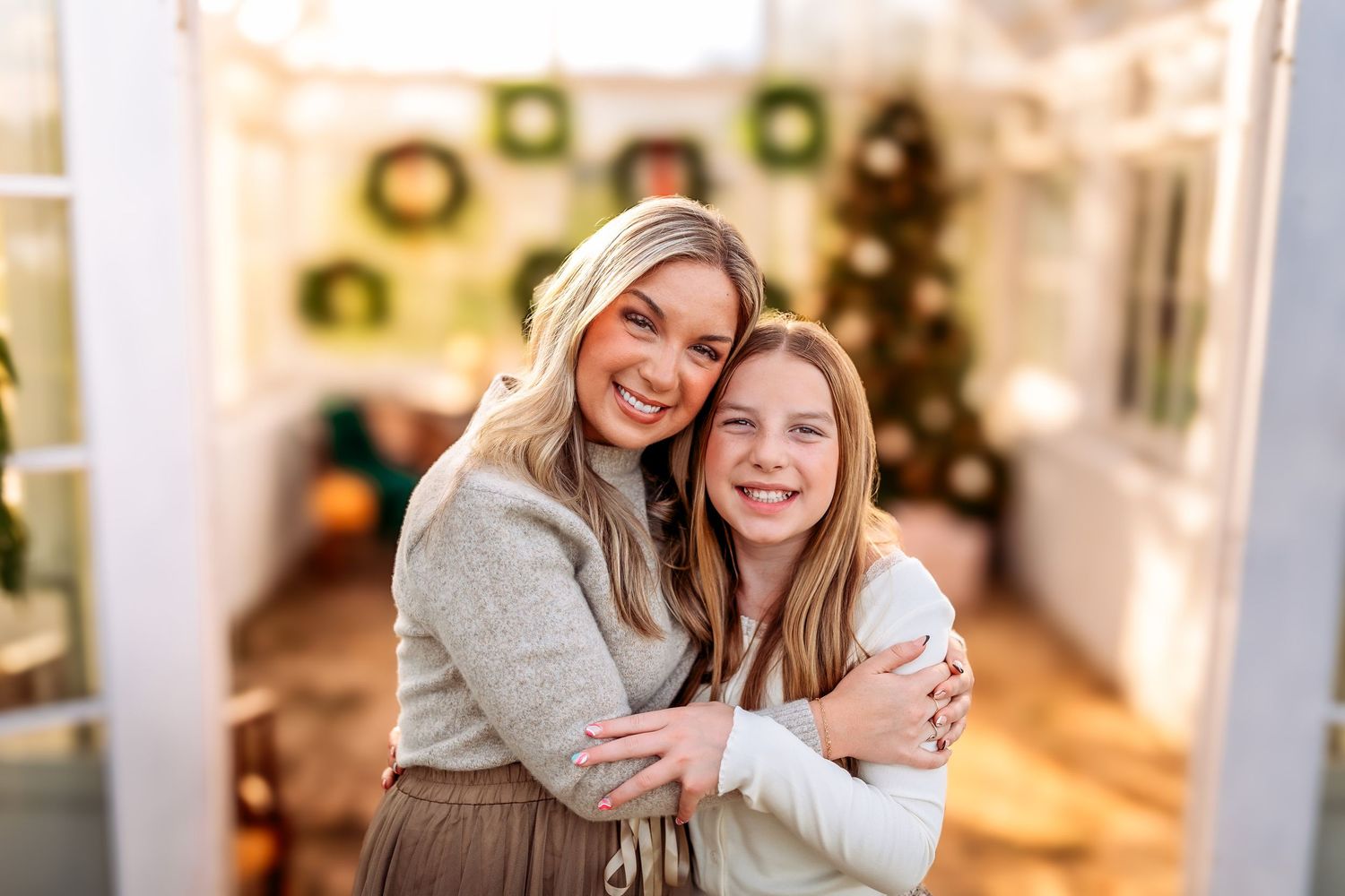 Two smiling women hugging in front of a decorated Christmas tree in a warmly lit holiday setting.