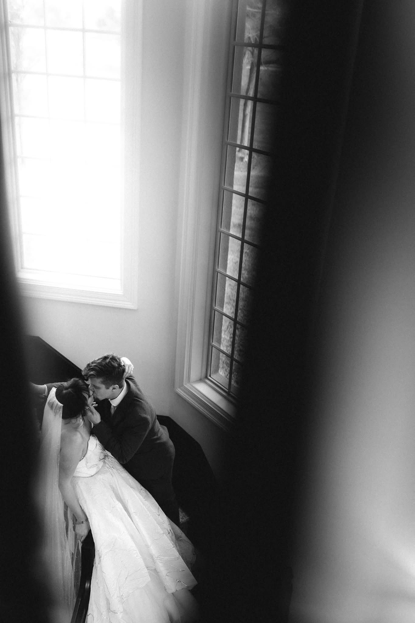 Black and white photo of a bride and groom sharing an intimate moment by large windows on their wedding day.
