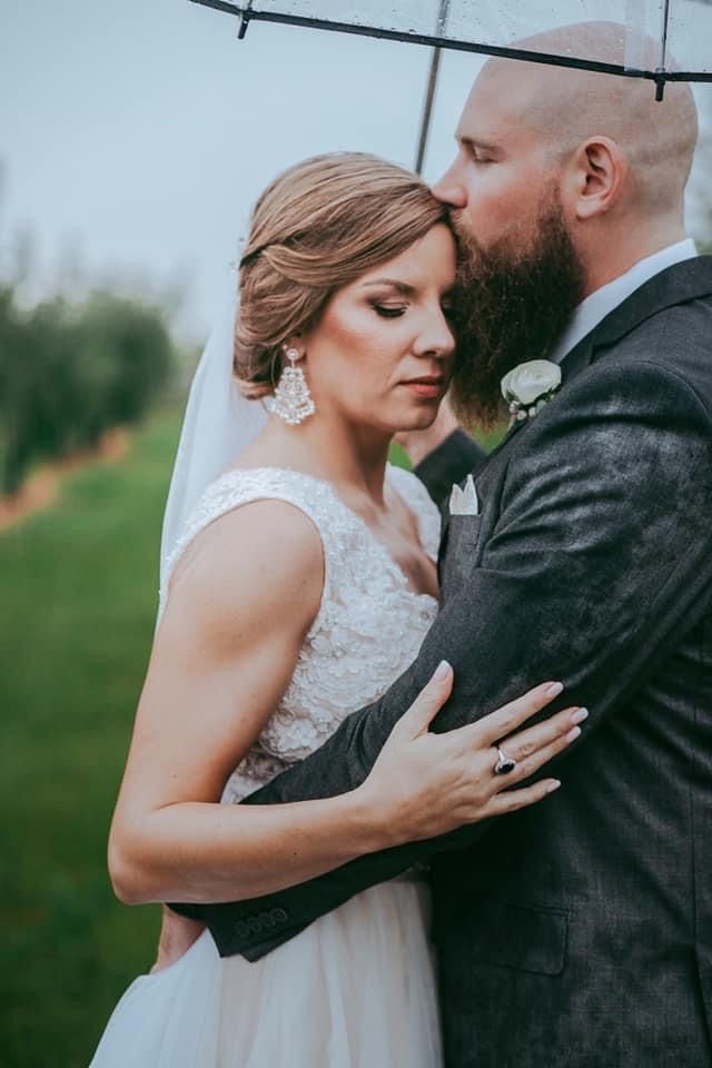 Bride and groom share tender moment under clear umbrella, groom kissing bride's forehead in lush green outdoor setting.