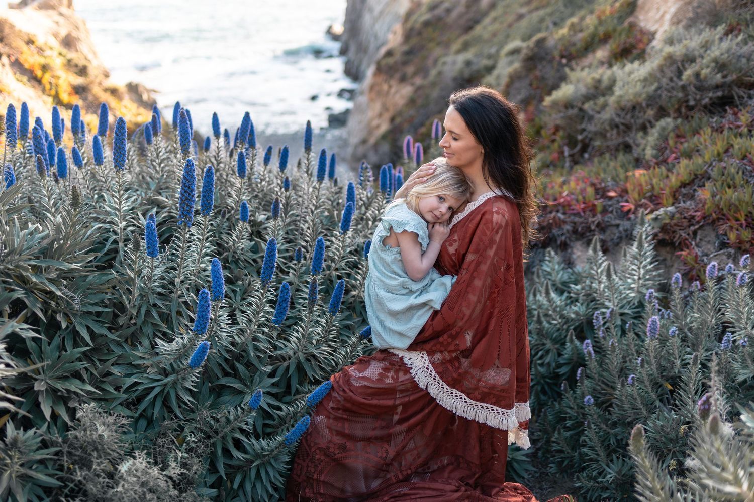Woman in red dress holds child among blue Pride of Madeira flowers on a coastal cliffside.