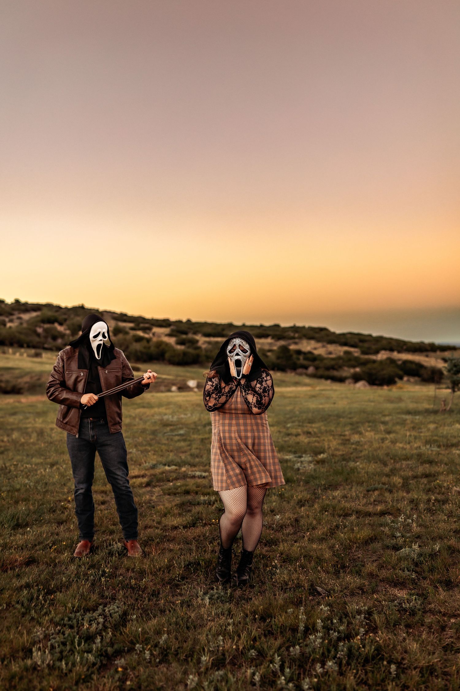 Two people in Ghostface masks pose in a grassy field at sunset, one holding a prop knife and the other clutching their face.