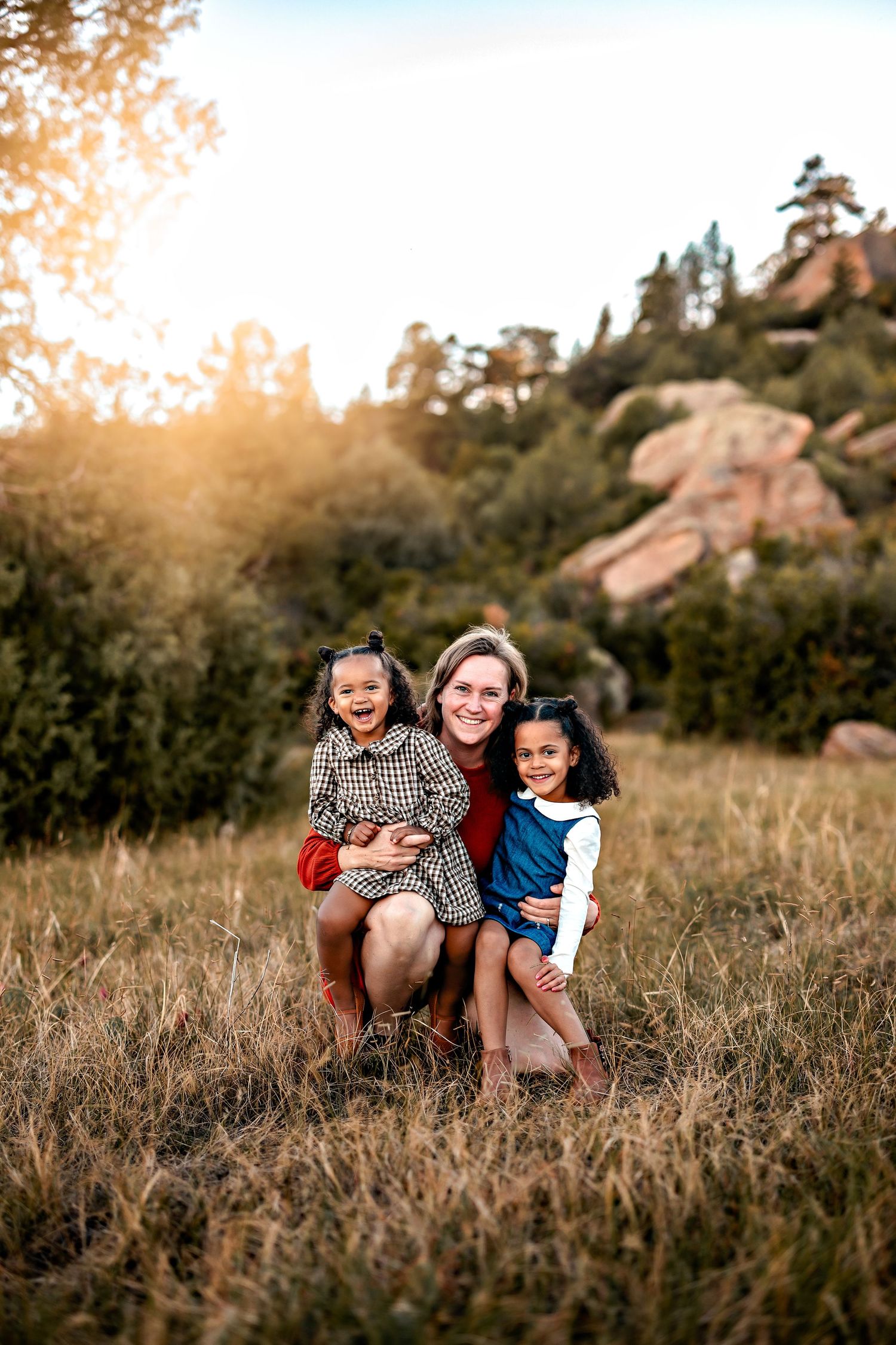 A smiling woman kneels in a sunlit grassy field hugging two giggling young girls, with rocky hills and trees glowing in the background.