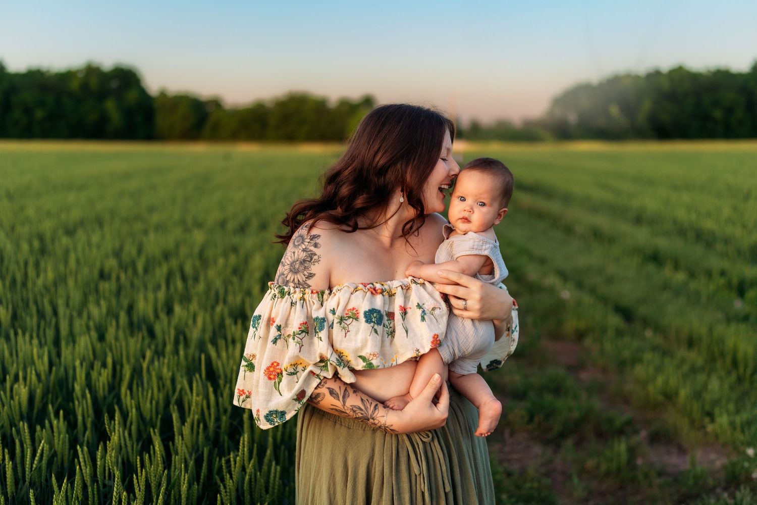 Mother in floral off-shoulder top holding baby in green field at soft golden hour sunset light.