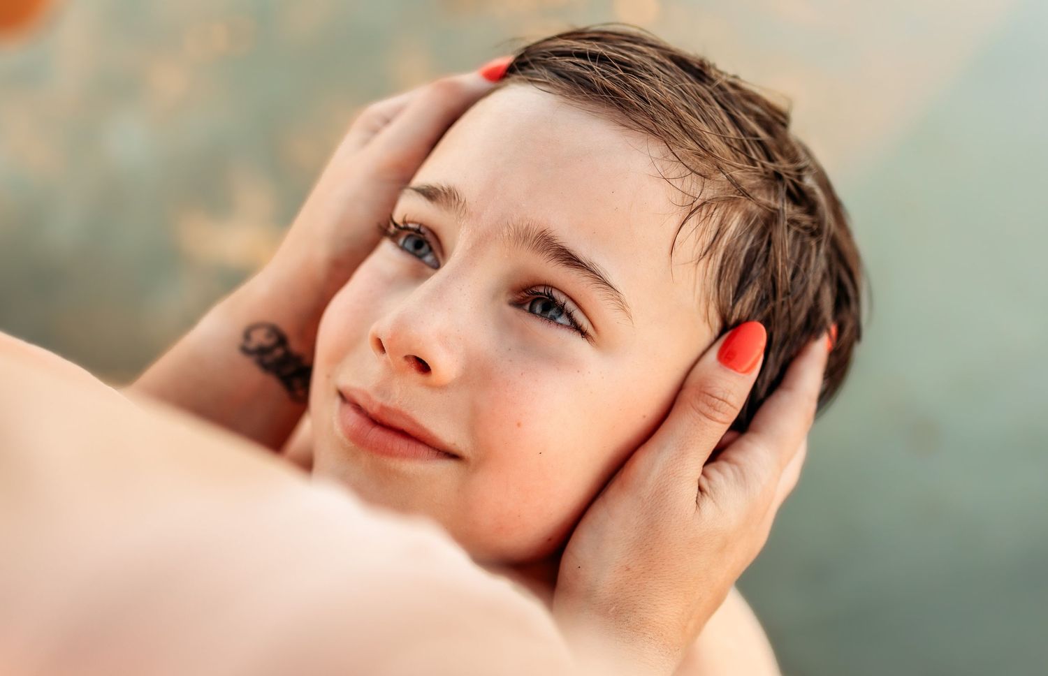 An adult tenderly holds a child's face in their hands with bright red nails, as the young boy looks up with a gentle smile.