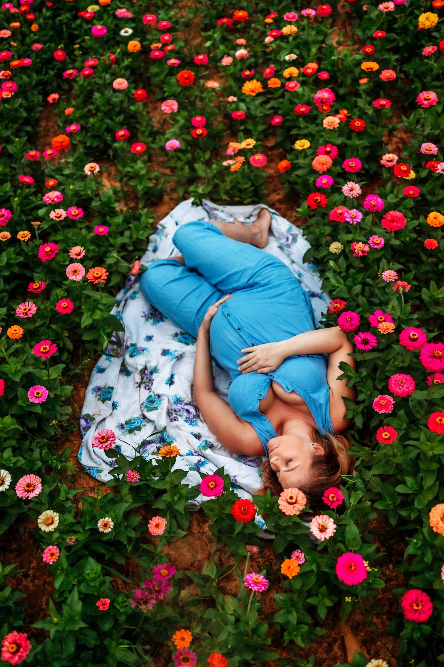 Pregnant woman in blue dress lying on floral blanket surrounded by colorful zinnia flowers in bloom.