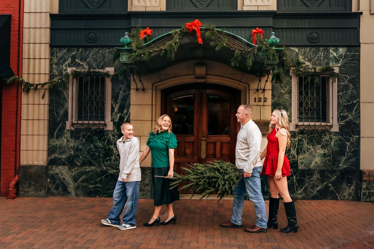 Family of four poses in front of a festive holiday-decorated building entrance on a brick sidewalk.