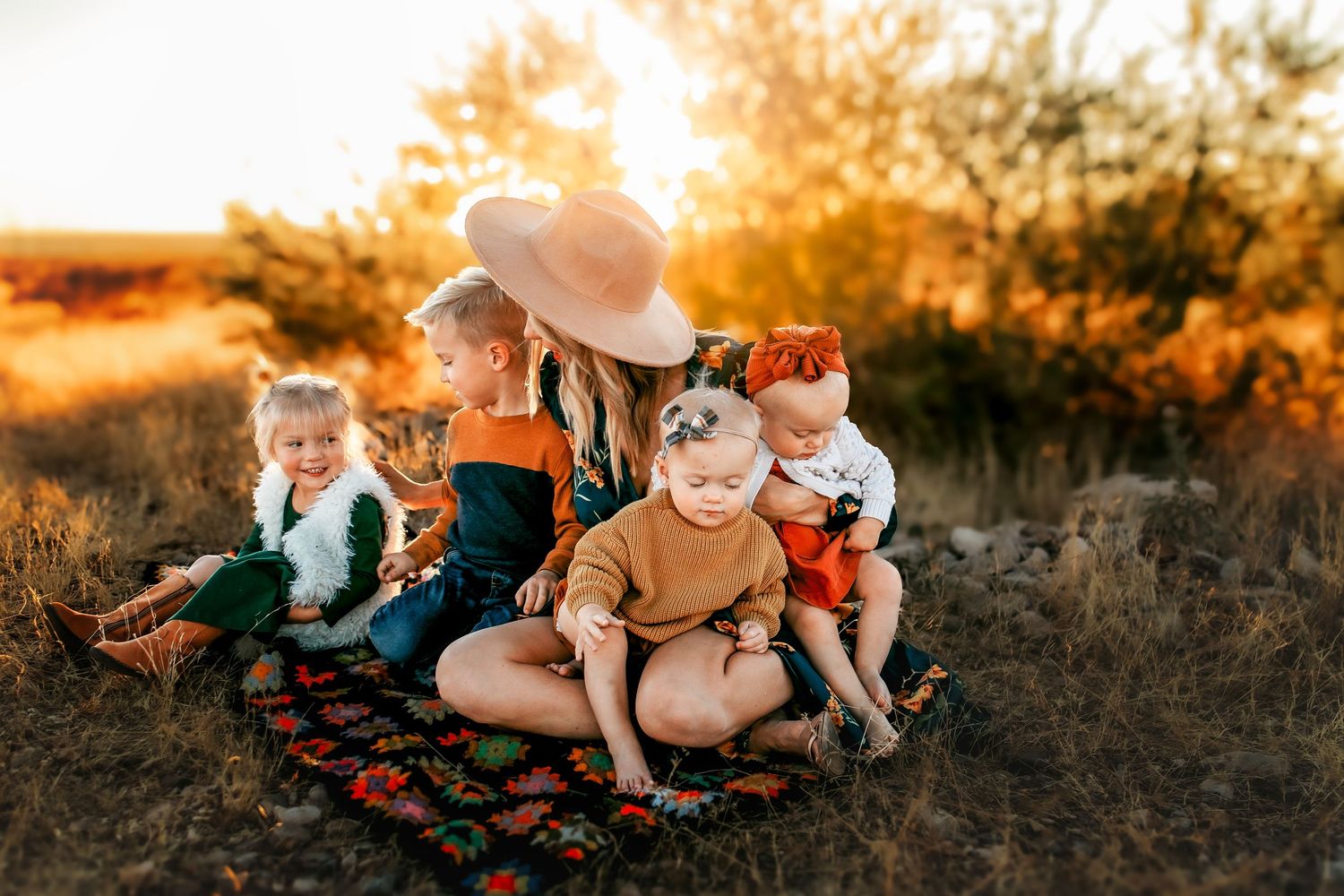 A mother in a wide-brim hat sits on a colorful blanket in a golden field with her four young children at sunset.