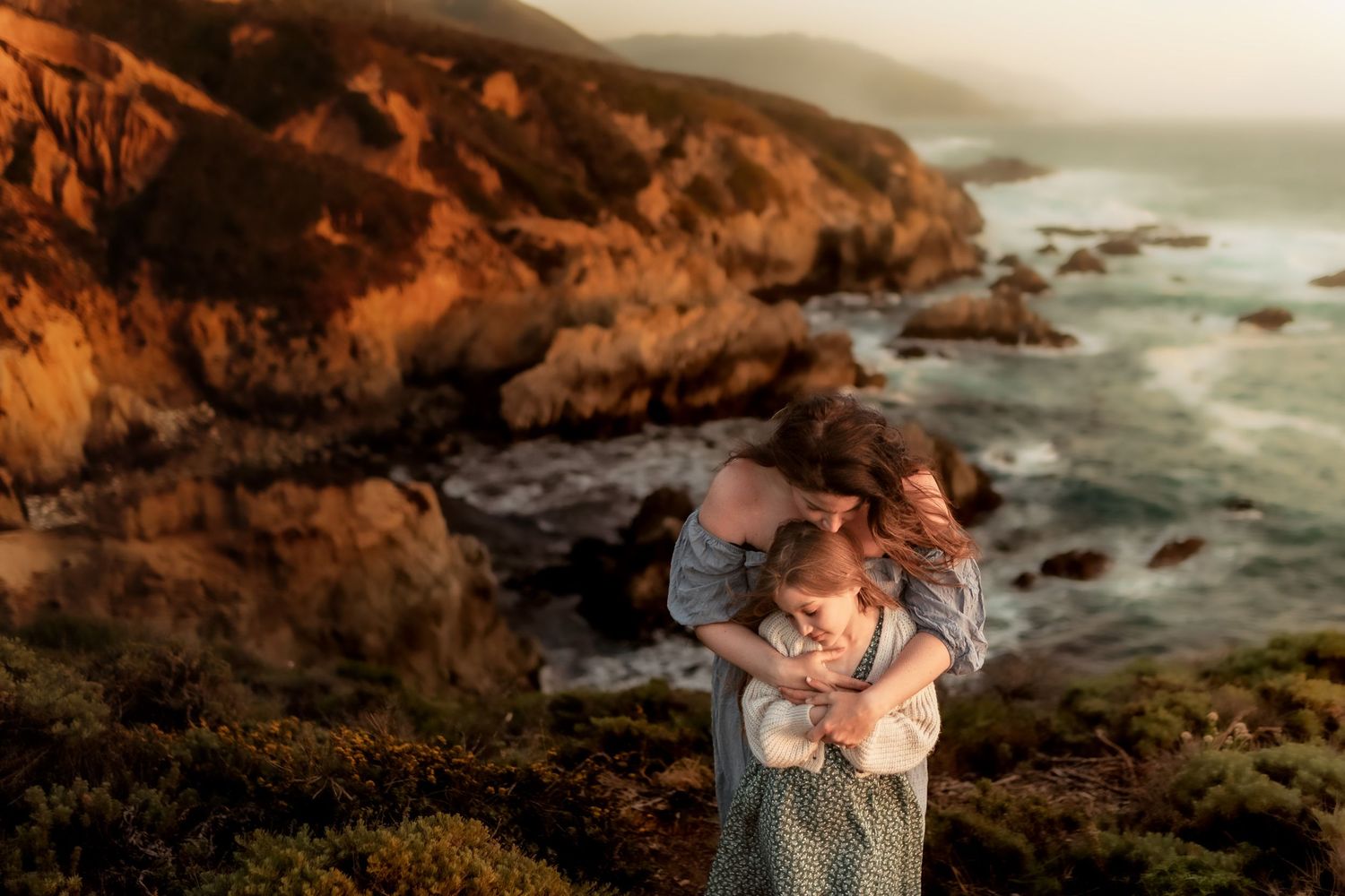 Mother and child embracing on a rocky Big Sur coastline at golden hour sunset.