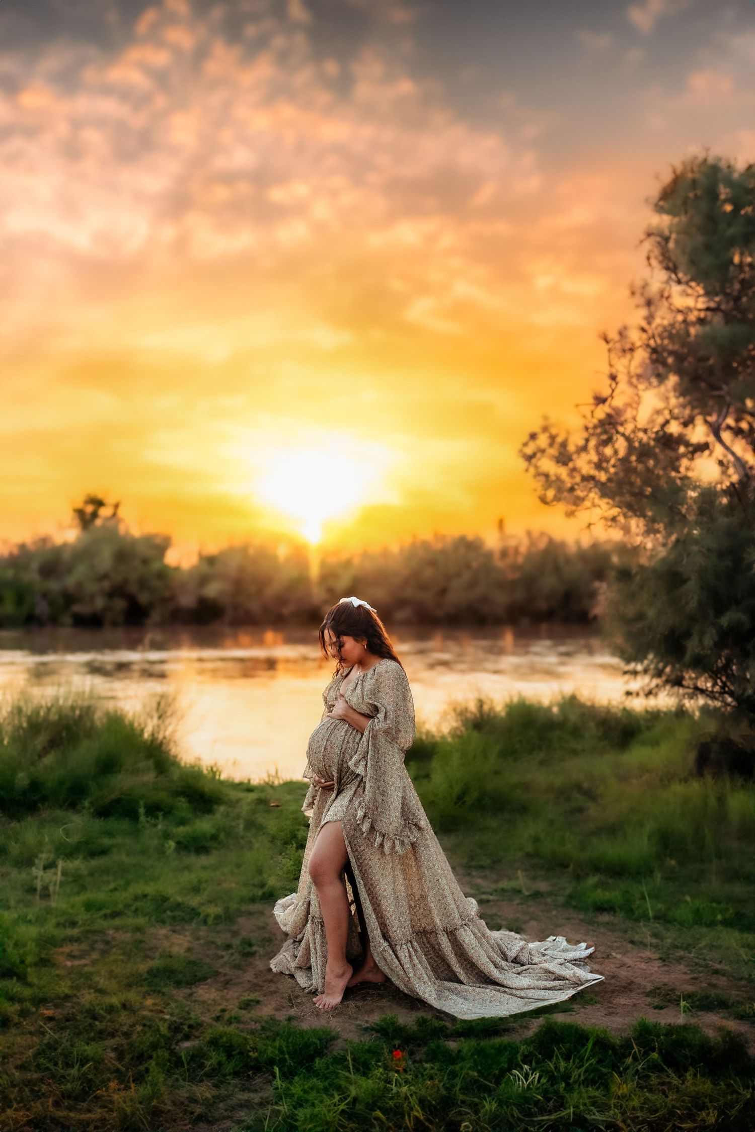 Pregnant woman in elegant lace gown standing by a river at golden sunset, cradling her baby bump.