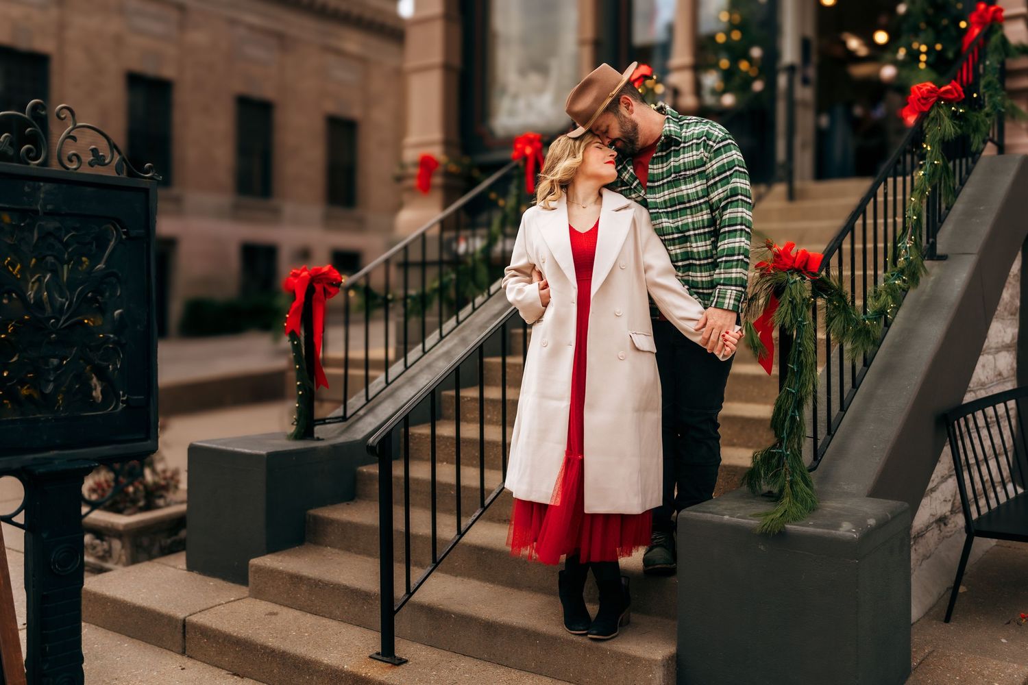 Couple holding hands on decorated holiday steps, woman in white coat and red dress, man in plaid jacket.