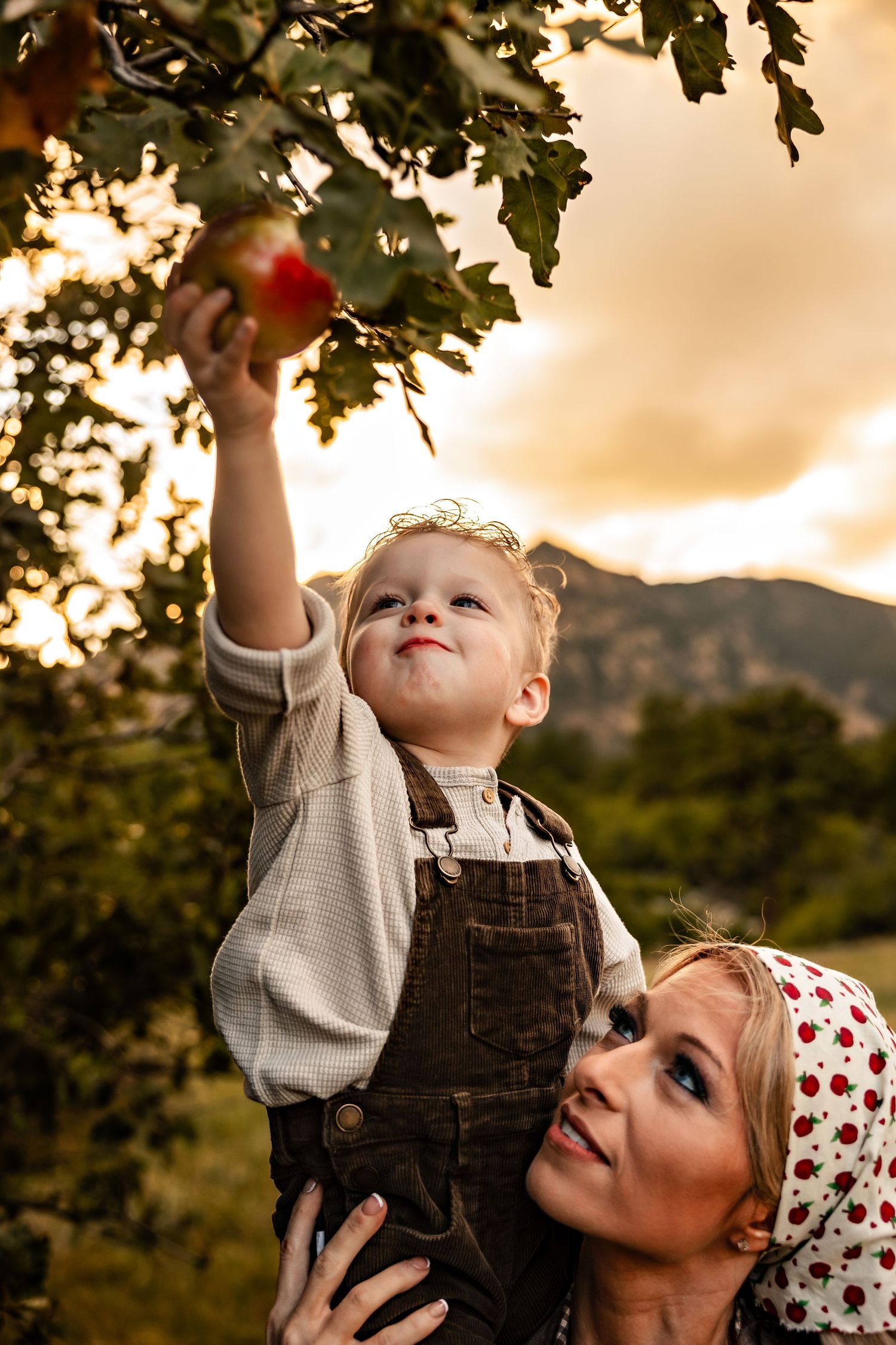 A woman in a polka dot headscarf lifts a toddler in overalls to reach a red apple on a tree, with mountains glowing at sunset behind them.
