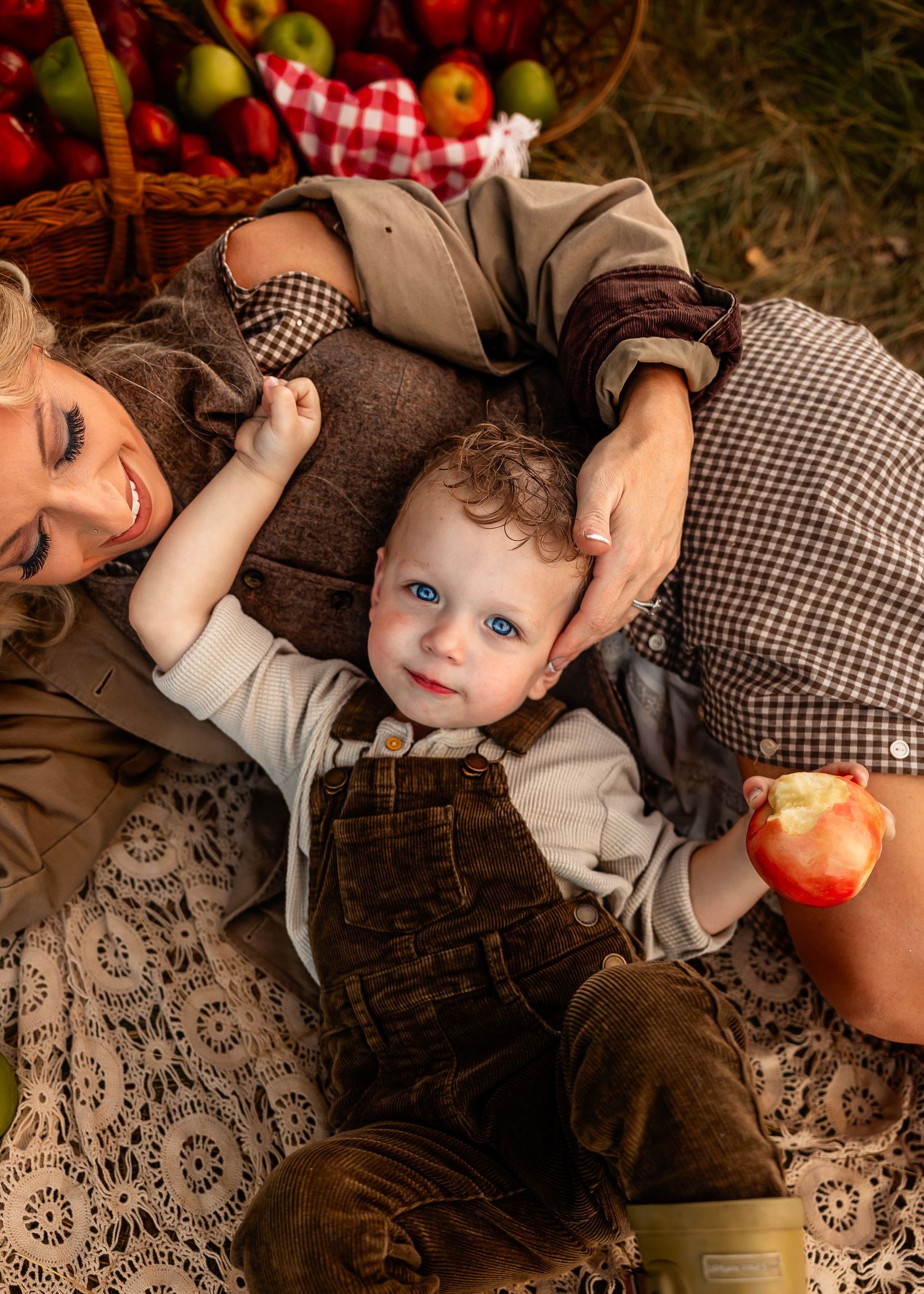 Smiling toddler in brown corduroy overalls holds a red apple while lying beside his mother on a lace blanket near a basket of apples.