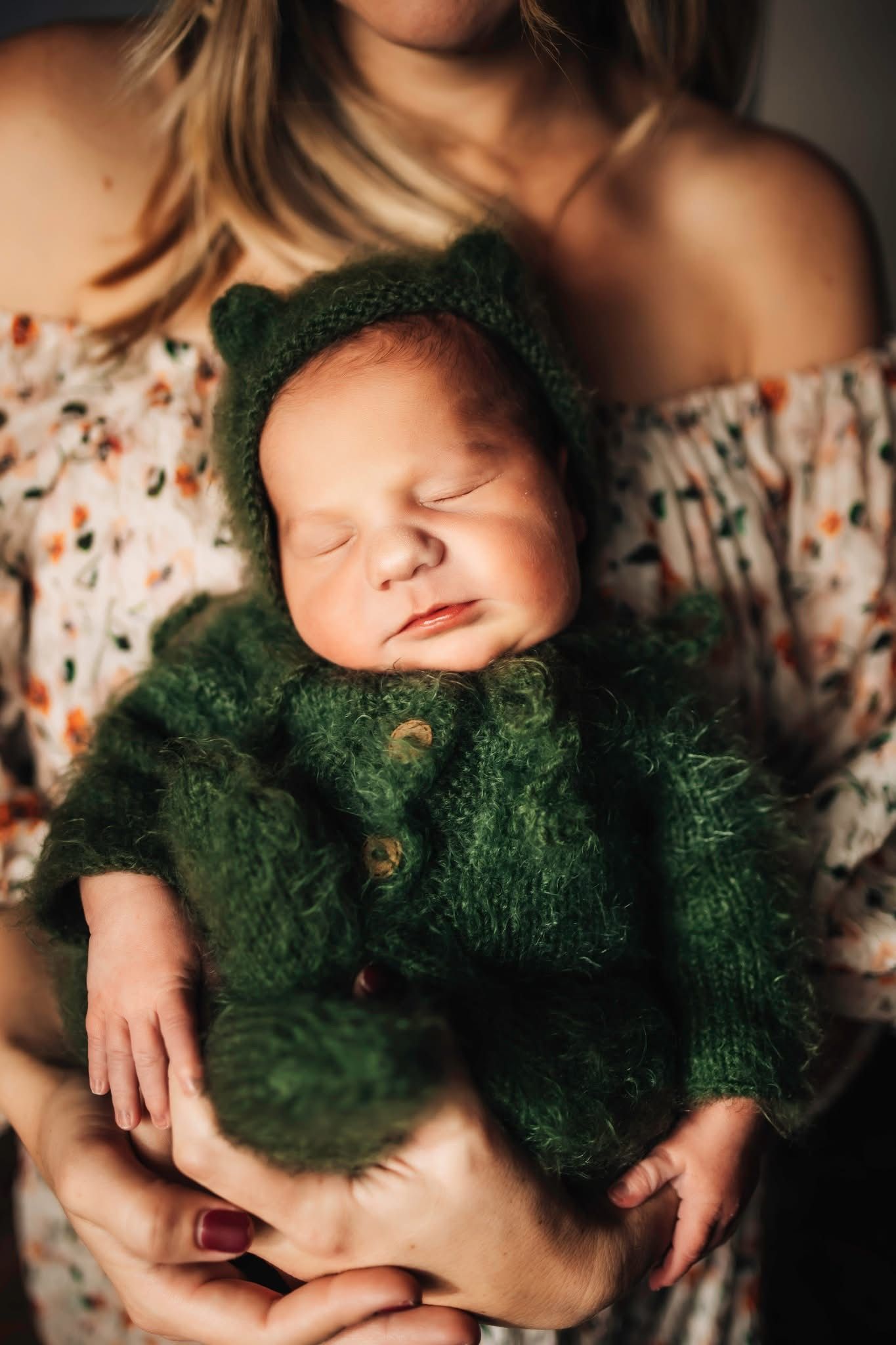 Newborn baby sleeping peacefully in green knit bear outfit, cradled in mother's hands.