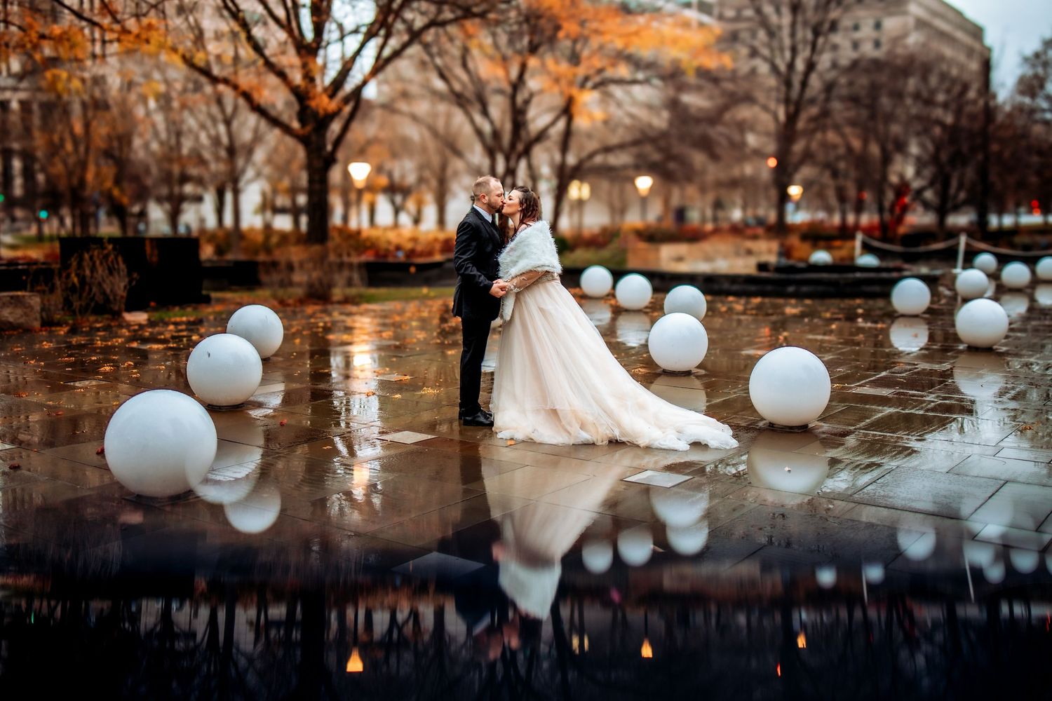 Bride and groom share a romantic kiss surrounded by white spheres on a reflective wet plaza with autumn trees.
