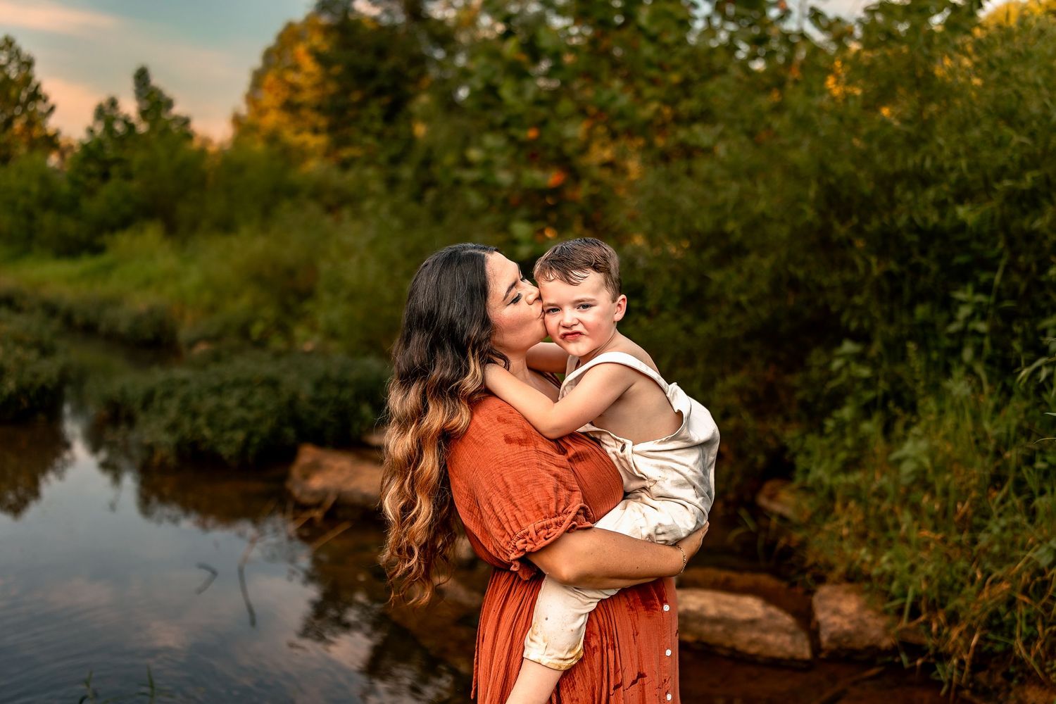 Mother in red dress kisses toddler by a sunlit creek surrounded by lush green trees at golden hour.