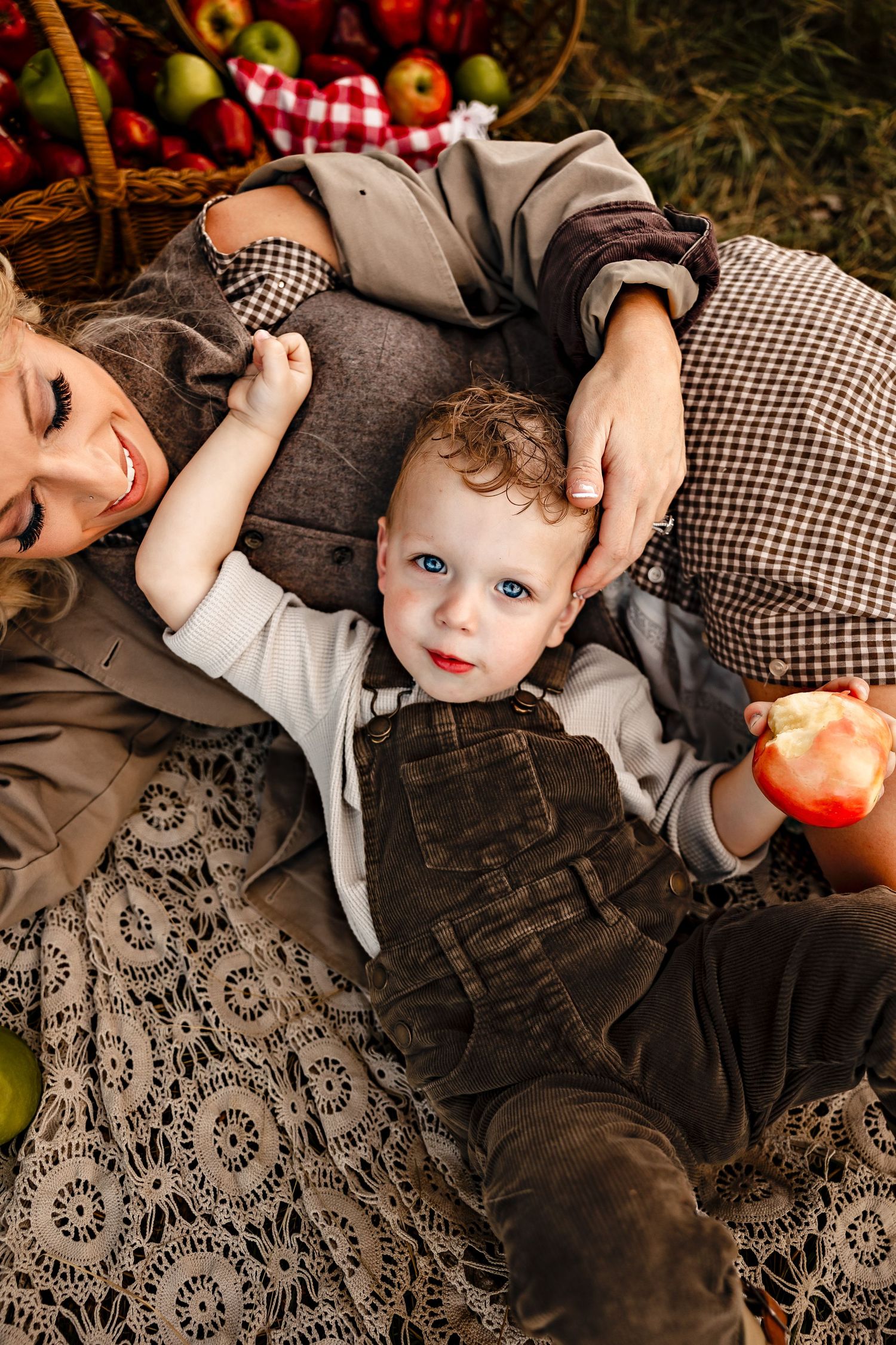 Smiling mother lying on a lace blanket holds a curly-haired toddler in corduroy overalls eating an apple beside a basket of fresh apples.