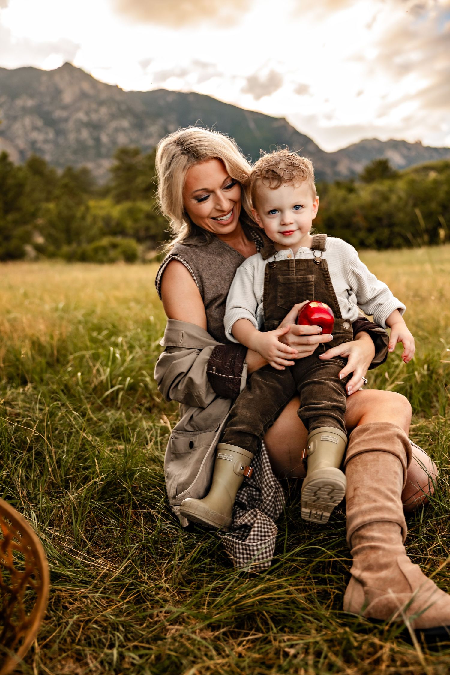 Smiling blonde mother in boots sits in a grassy field with her toddler son holding a red apple, with mountains in the background.
