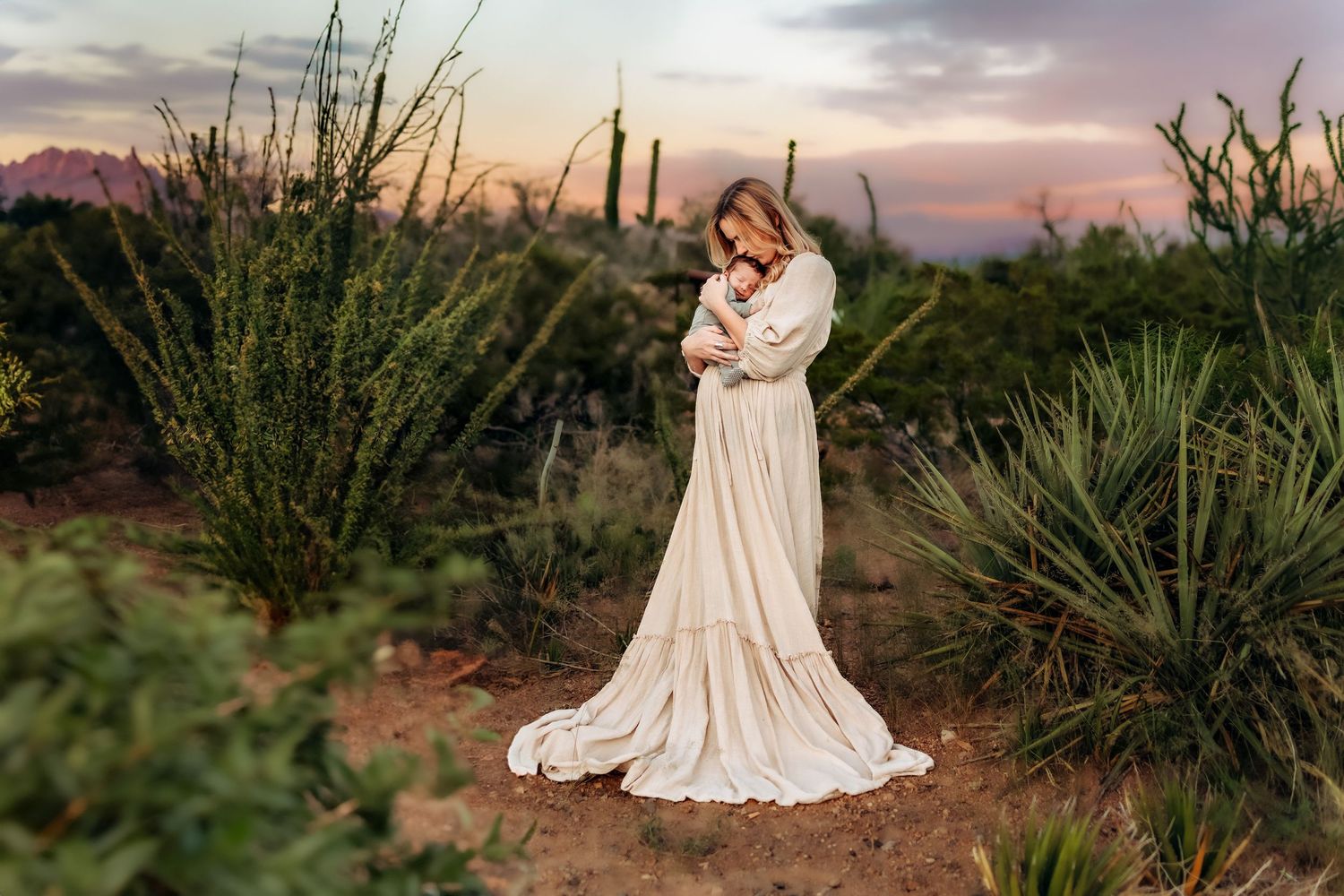A woman in a flowing cream gown holds a baby amid desert cacti at sunset in a serene maternity portrait.
