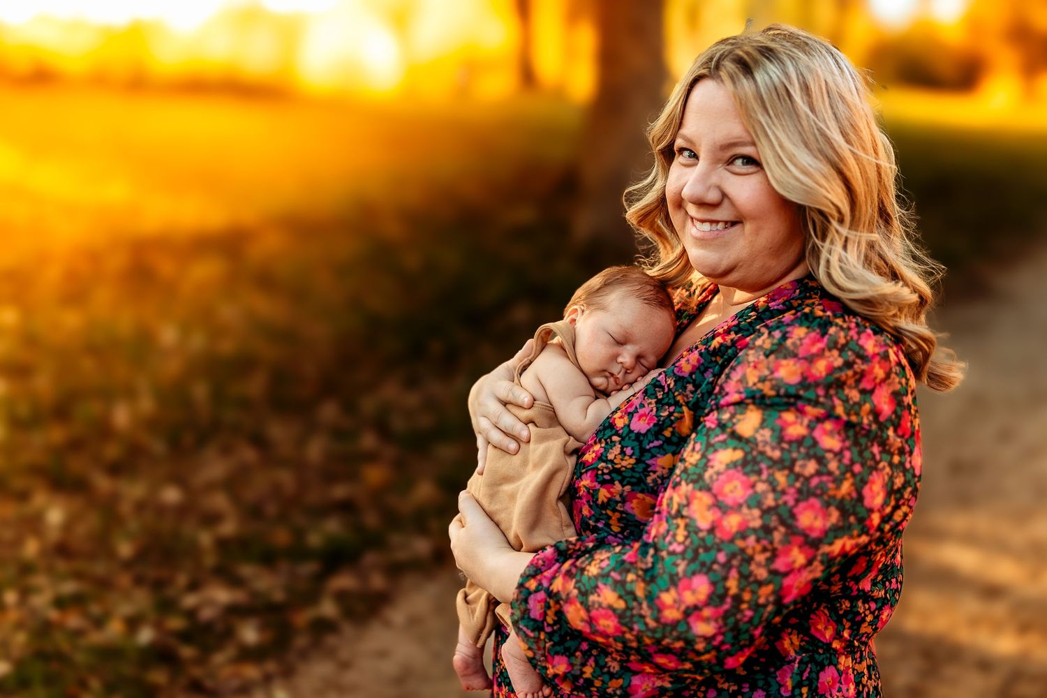 Smiling woman in floral dress holding sleeping newborn baby outdoors during golden sunset in autumn park.