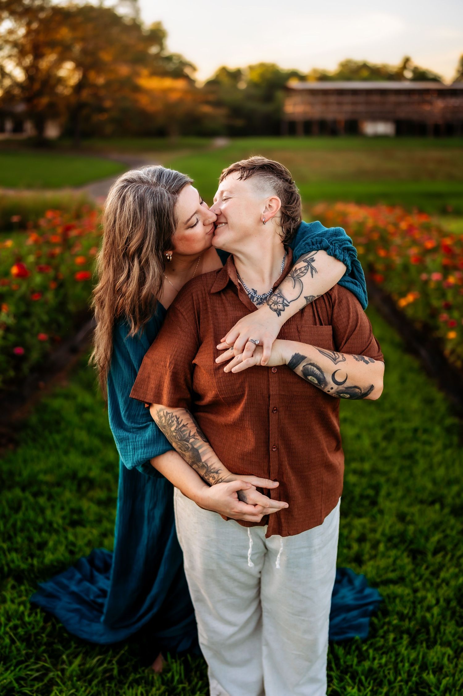 Two tattooed women share a tender kiss in a lush garden at golden hour, one in teal dress, other in rust shirt.