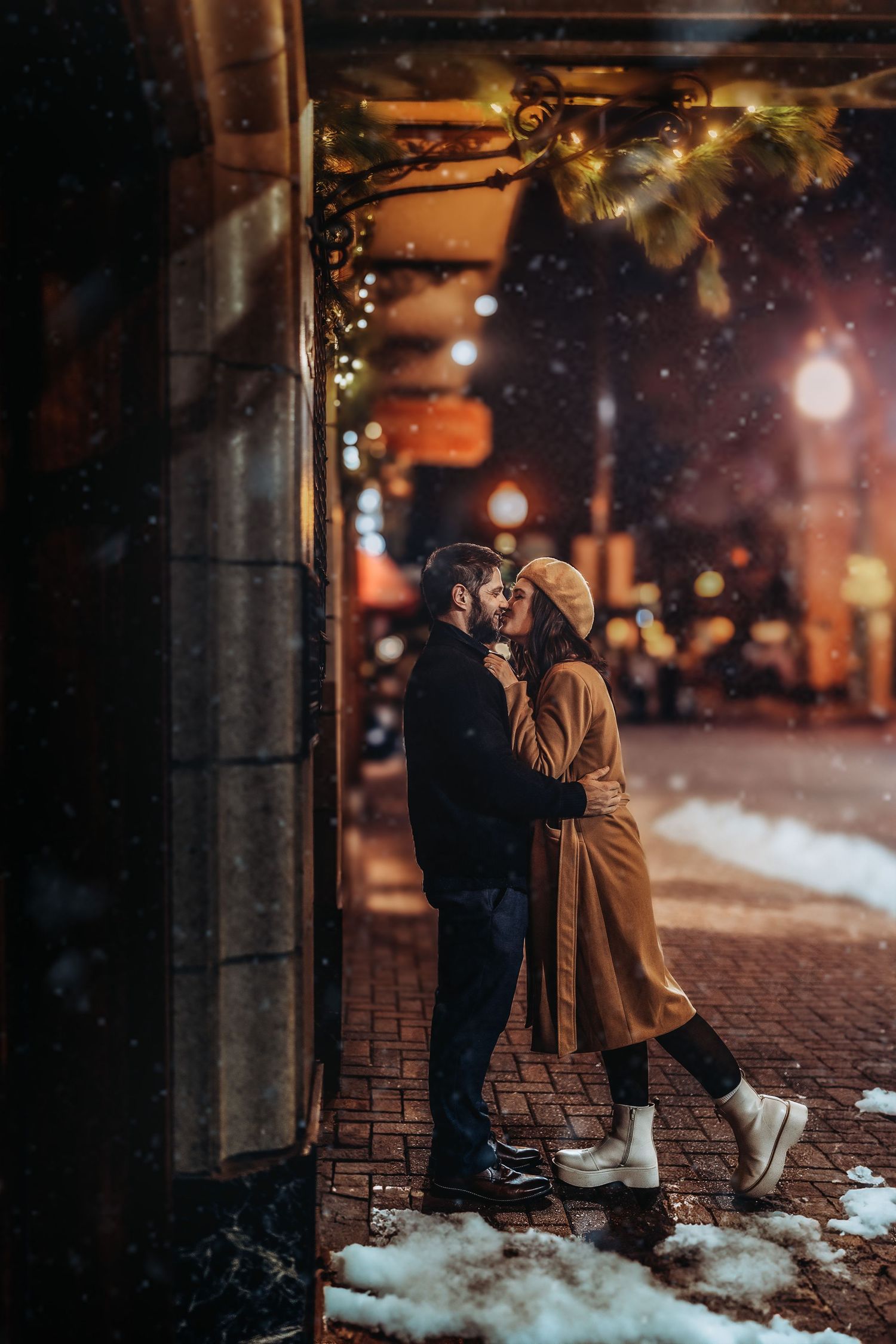 Couple sharing a romantic kiss in snowy city street at night, decorated with festive lights and greenery.