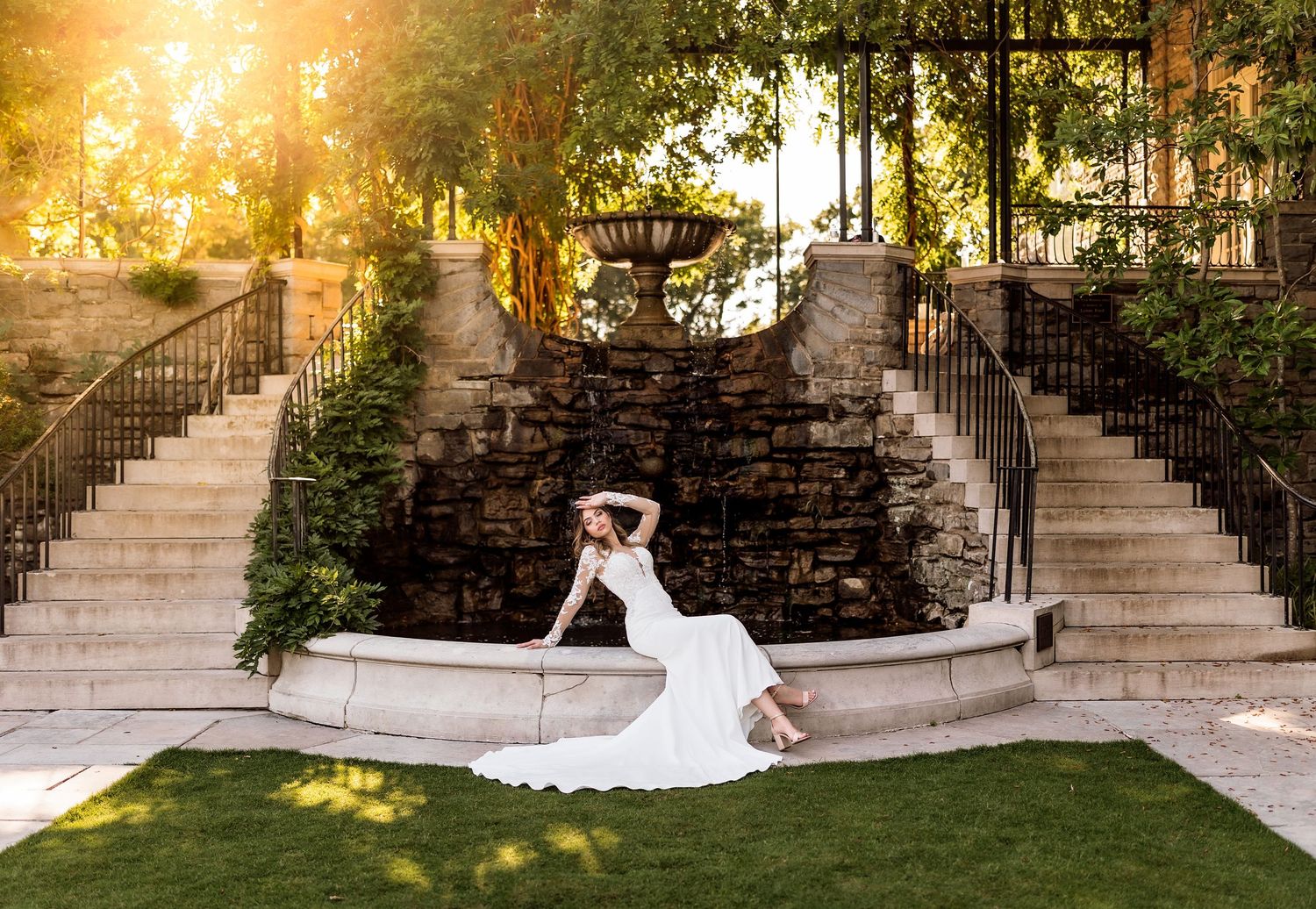 Bride in white gown posing elegantly by a stone fountain with cascading water, lush greenery, and golden sunlight.