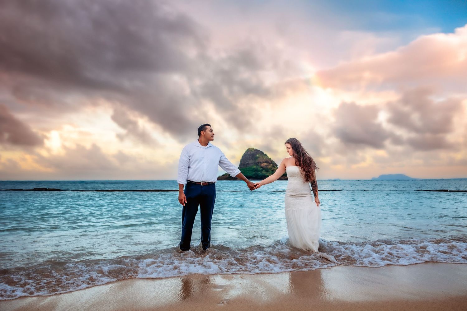 Couple holds a heart-shaped rock on a tropical beach at sunset with dramatic clouds and rainbow in the background.