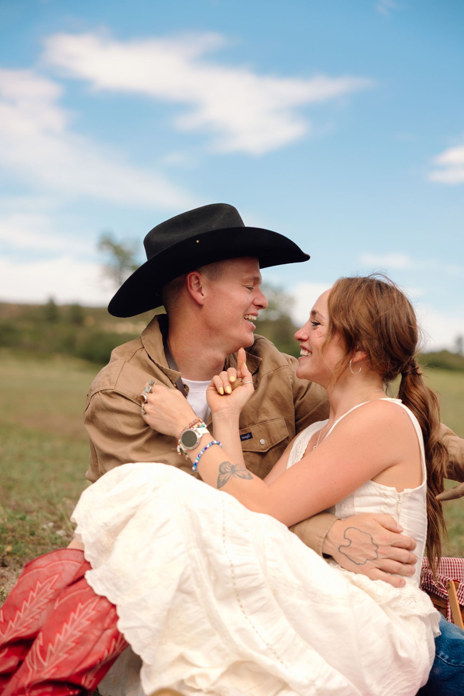 A smiling couple sits outdoors, man in black cowboy hat holds woman in white dress on his lap in a green field.