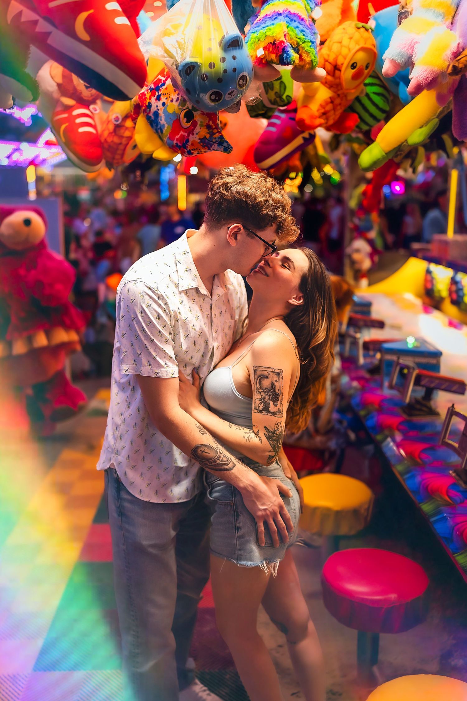 Couple kissing at a colorful carnival game booth surrounded by stuffed animal prizes and vibrant neon lights.