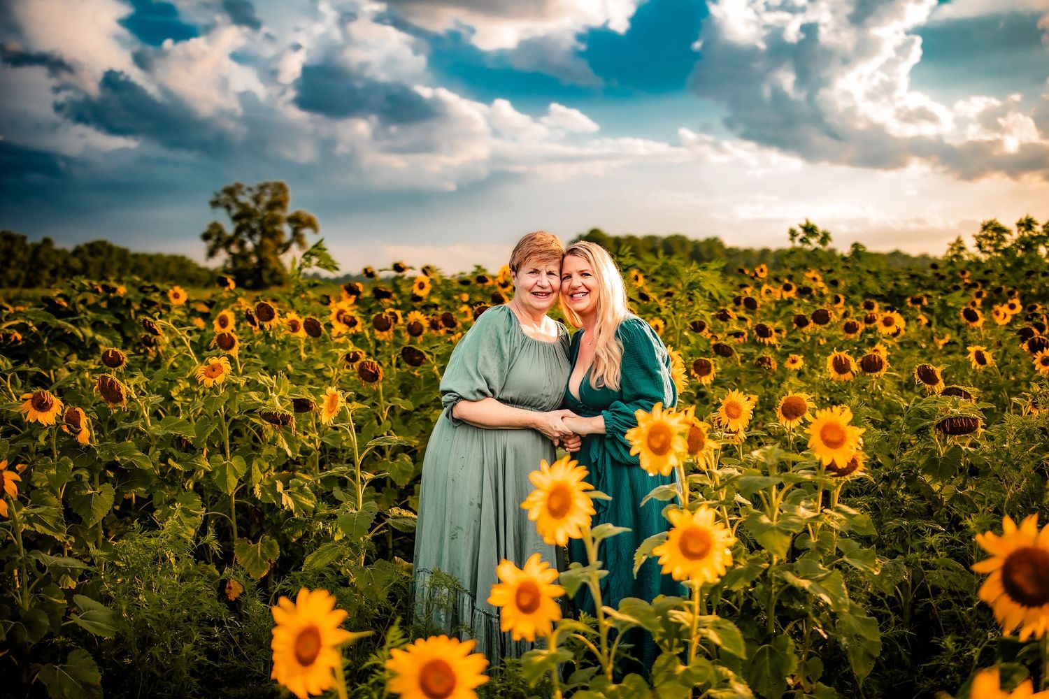 Two women in teal dresses embrace in a vibrant sunflower field under a dramatic cloudy sky at sunset.