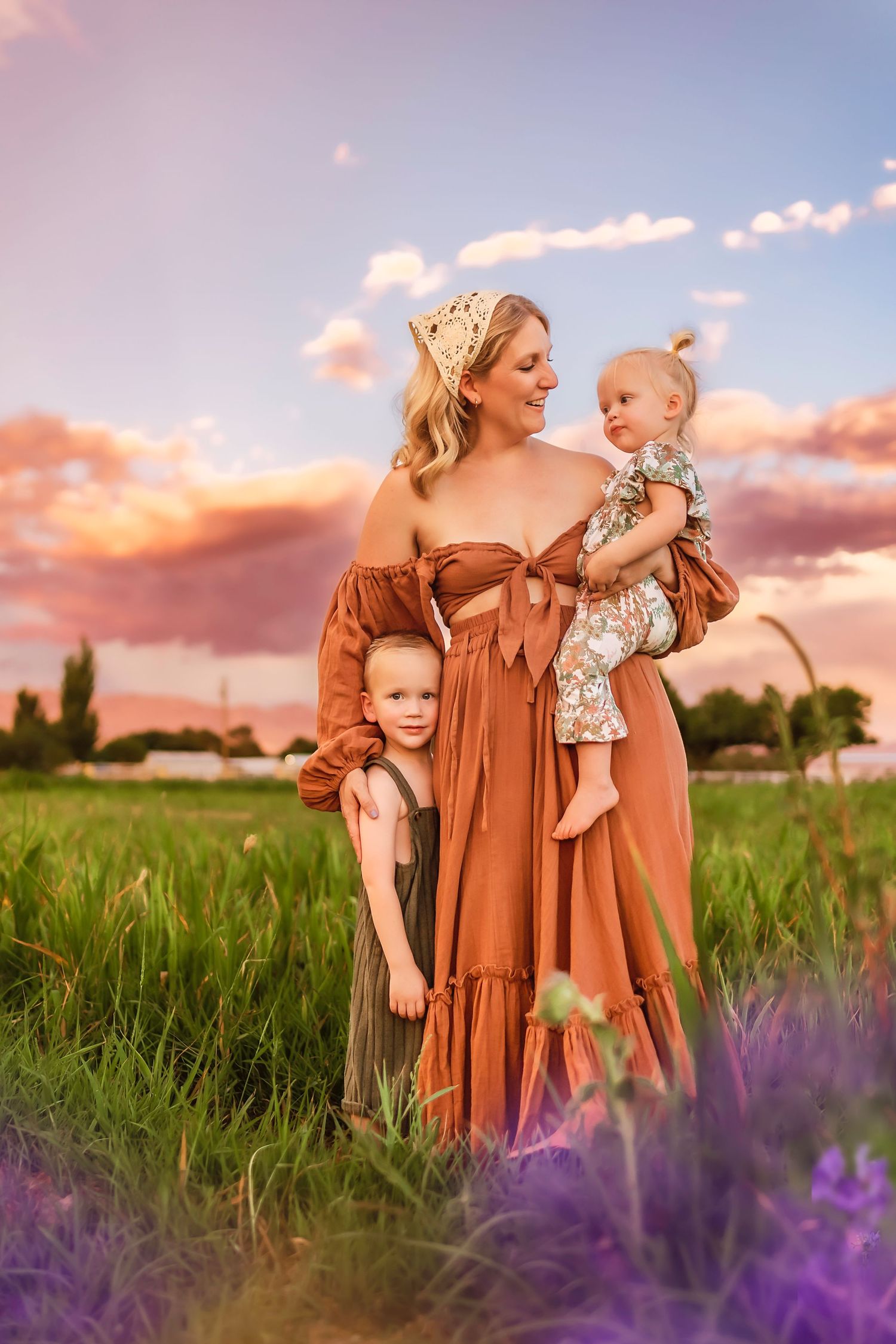Mother in rust dress holds baby girl while young boy hugs her in a golden sunset field.