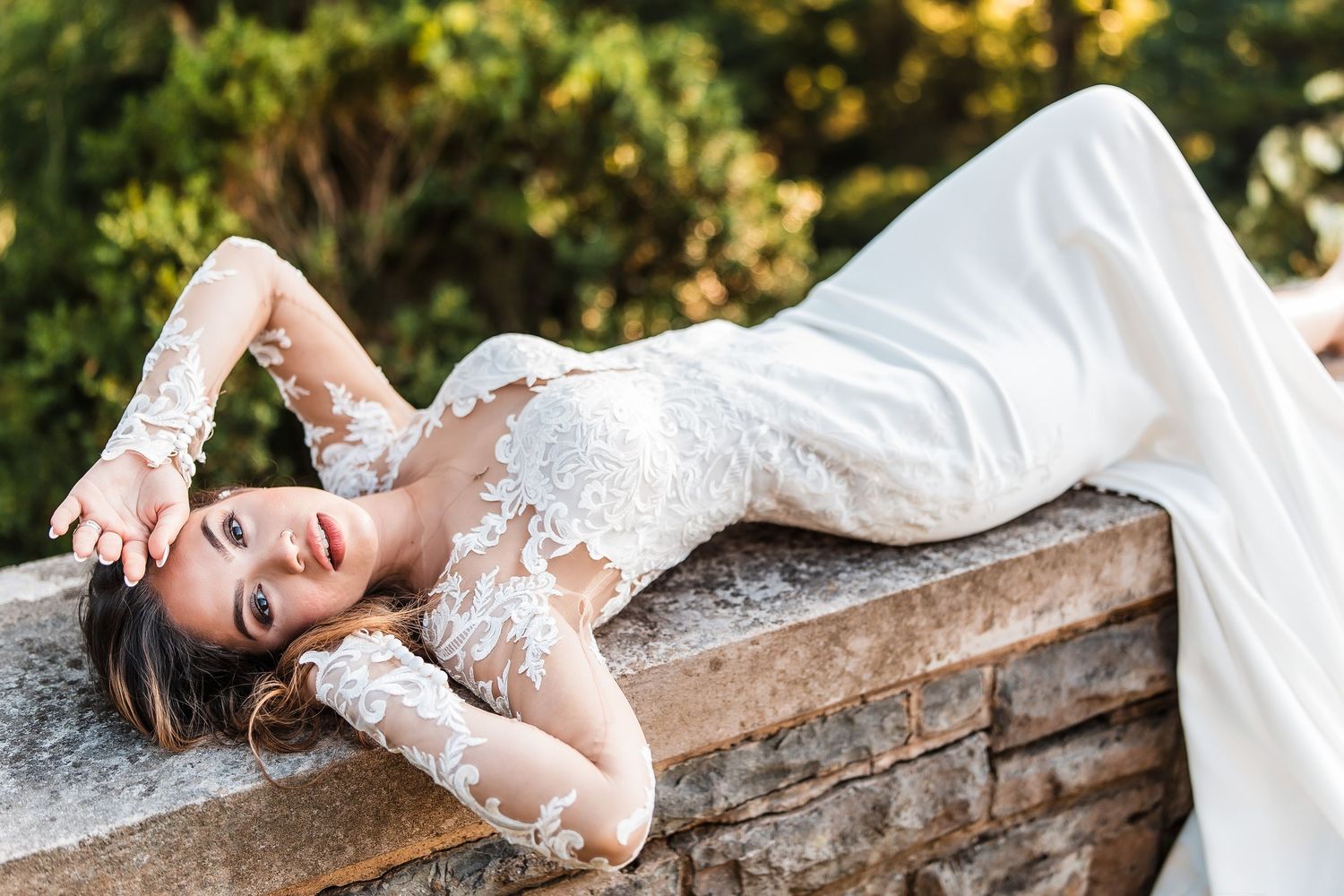 Bride in lace long-sleeve wedding gown lying gracefully on stone wall surrounded by lush greenery.