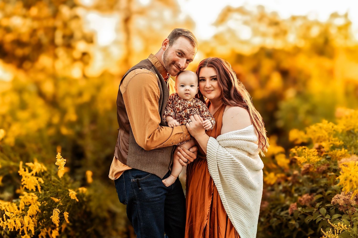 Family of three posing outdoors in autumn foliage, parents holding baby in fall-themed outfits during golden hour.