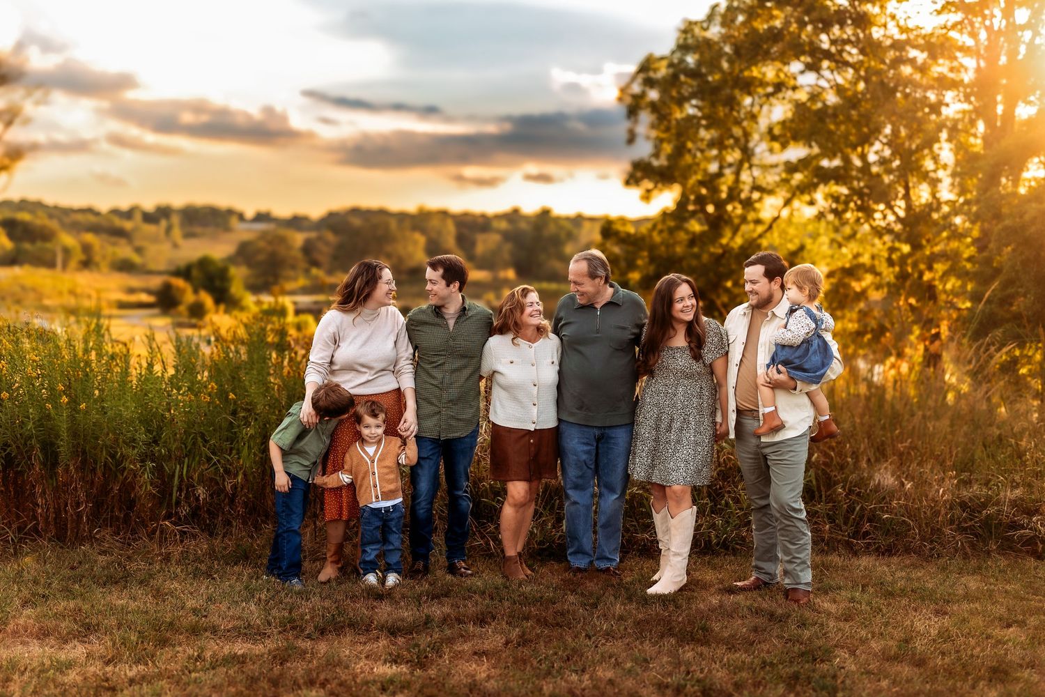 Family portrait at golden hour in a field, seven people smiling together with warm sunset light and trees in background.