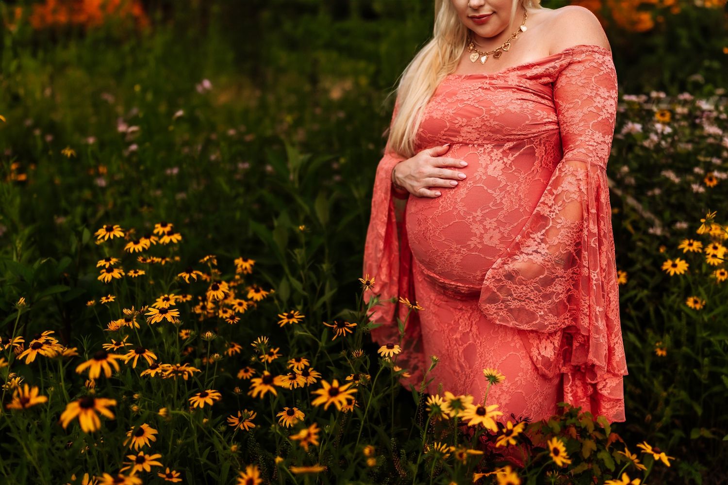 Pregnant woman in red lace off-shoulder gown cradling baby bump among yellow wildflowers in a garden.