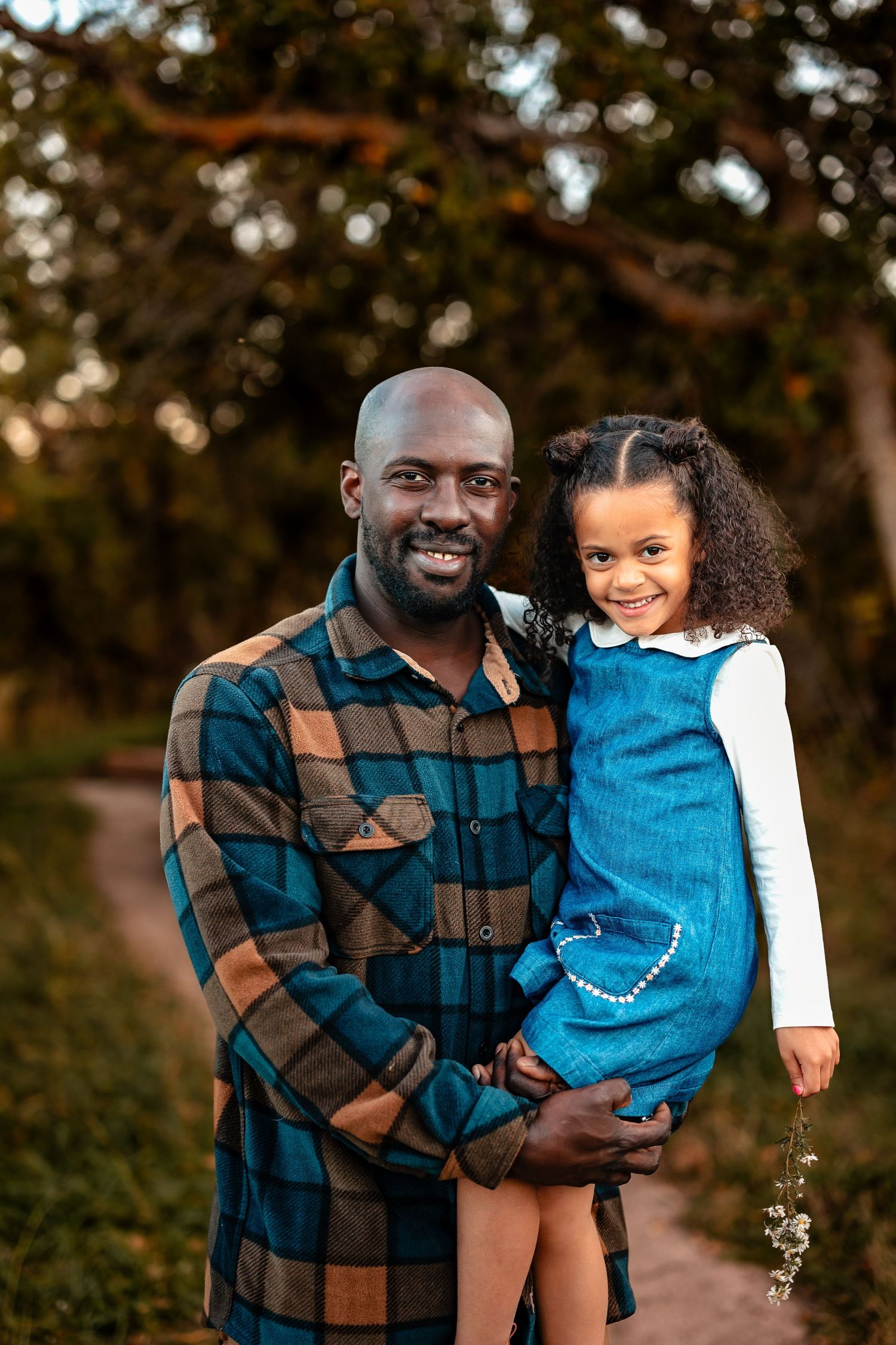 Smiling man in a plaid flannel shirt holding a happy young girl in a denim dress on a tree-lined outdoor path at golden hour.