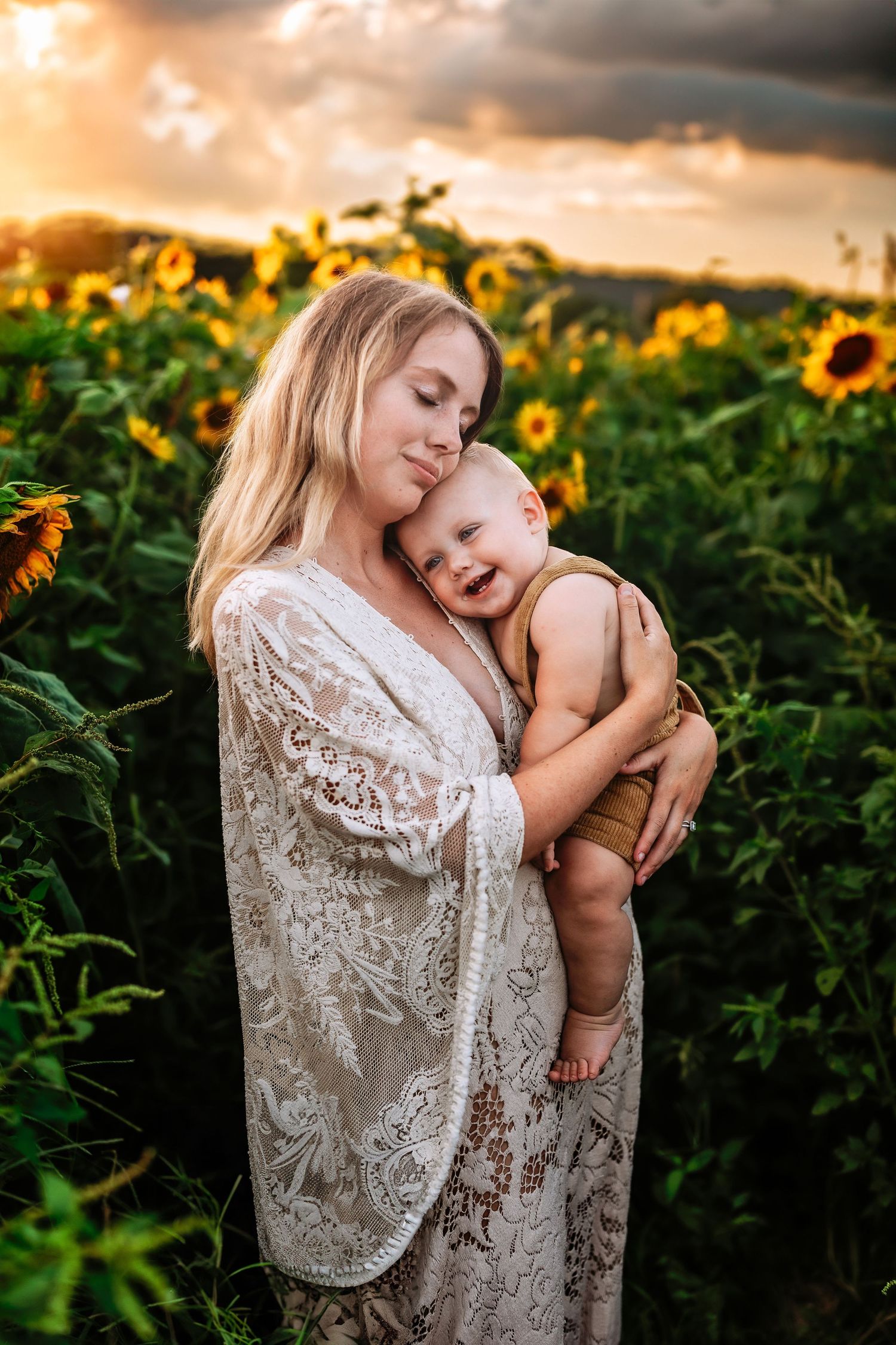 Mother in white lace dress holding laughing baby in sunflower field at golden sunset.