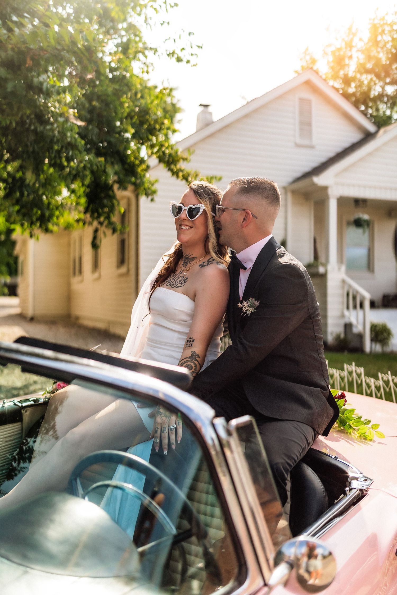 Bride and groom sitting on classic convertible car, sharing a kiss, bride wearing heart sunglasses, sunny outdoor wedding.