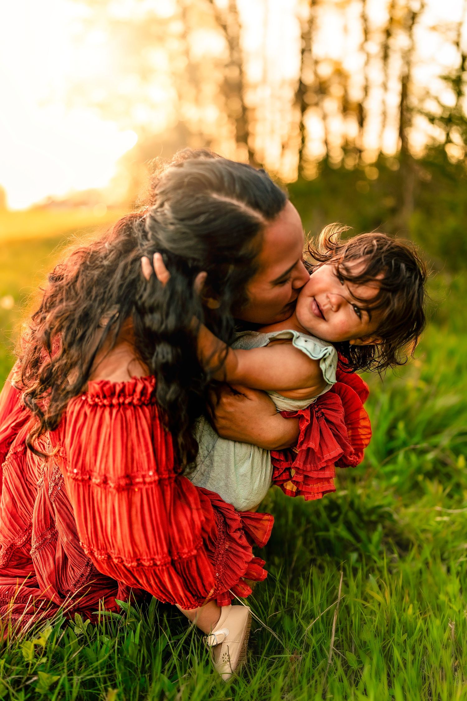 Mother in red dress kisses smiling toddler in sunny green field at golden hour.