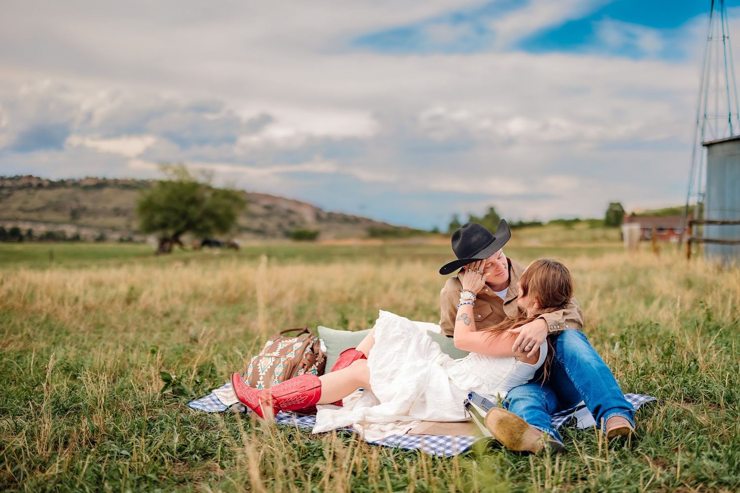 Couple relaxing on a picnic blanket in a scenic rural field, sharing a romantic moment under a cloudy sky.