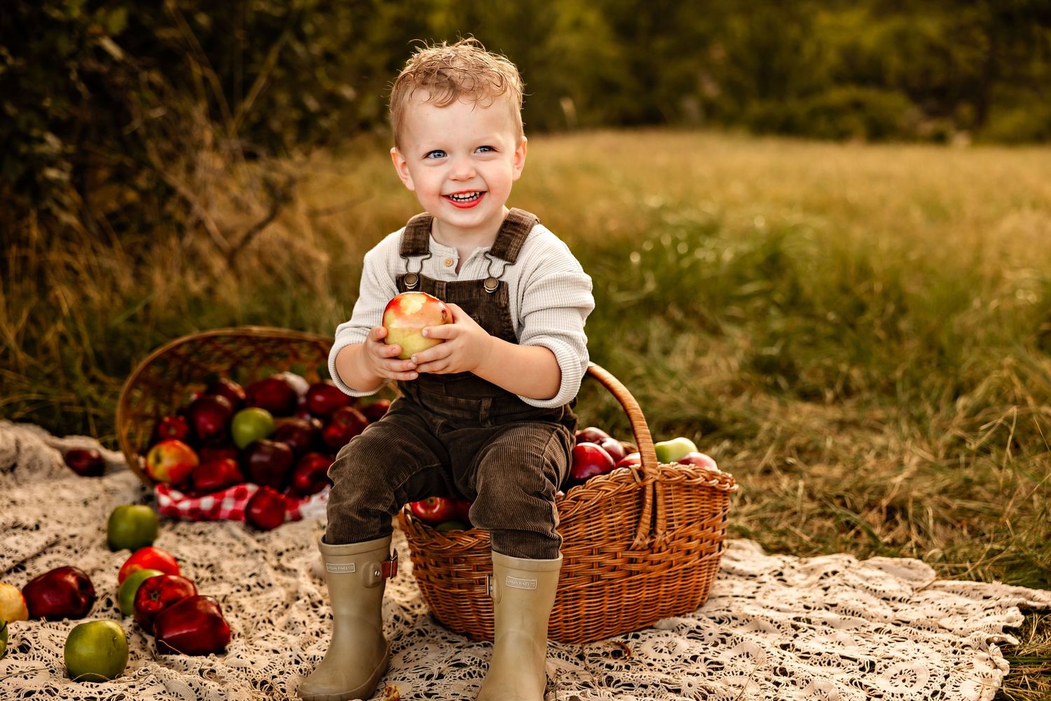 Smiling toddler in overalls and rubber boots sits on a wicker basket holding a red apple on a blanket in an autumn orchard.