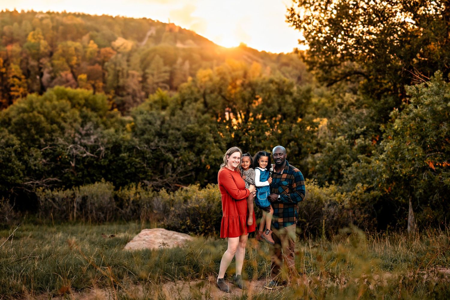 Family of four in a grassy field at golden hour, with a woman in a red dress, a man holding a child, and autumn trees behind.